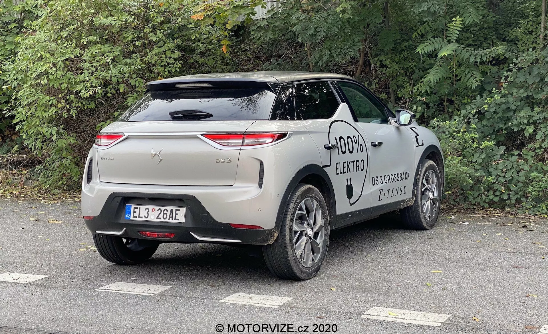DS Automobiles DS 3 2019 (DS 3 Crossback E-Tense) posnet z zadnjega tričetrtinskega kota, poudarjajoč zadnji in bočni profil vozila. Oblikovne značilnosti vključujejo horizontalno usmerjene zadnje luči, kromirano obrobo, gladke ročaje vrat, polnilno odprtino in večkrake lita platišča, značilne za sodoben električni avto. Avto je parkiran na prostem v urbanem ali primestnem okolju pod oblačnim nebom, z listavci v bližini.