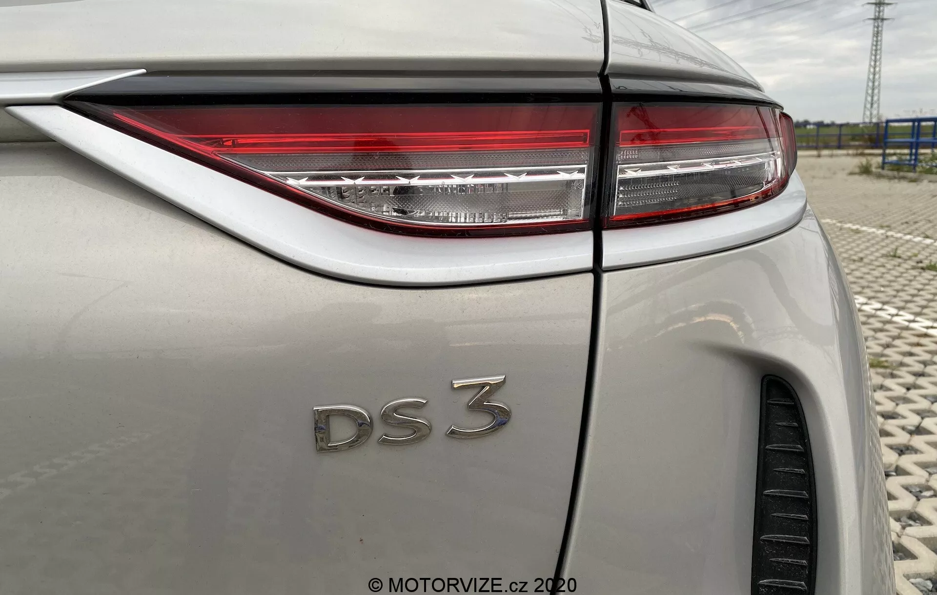 Bližnji pogled na zadnji trikotni kot DS Automobiles DS 3 Crossback E-Tense 2019. Slika se osredotoča na desno stran vozila, poudarjajoč ostre LED zadnje luči, značko 'DS 3' in lahko kovinsko karoserijo. Vidne lastnosti vključujejo linijo pregiba zadnjega blatnika, delno vidno zadnjo pnevmatiko, anteno v obliki plavuti morskega psa in prehod zadnjega stekla v linijo strehe, vse pod odsevom oblačnega neba.