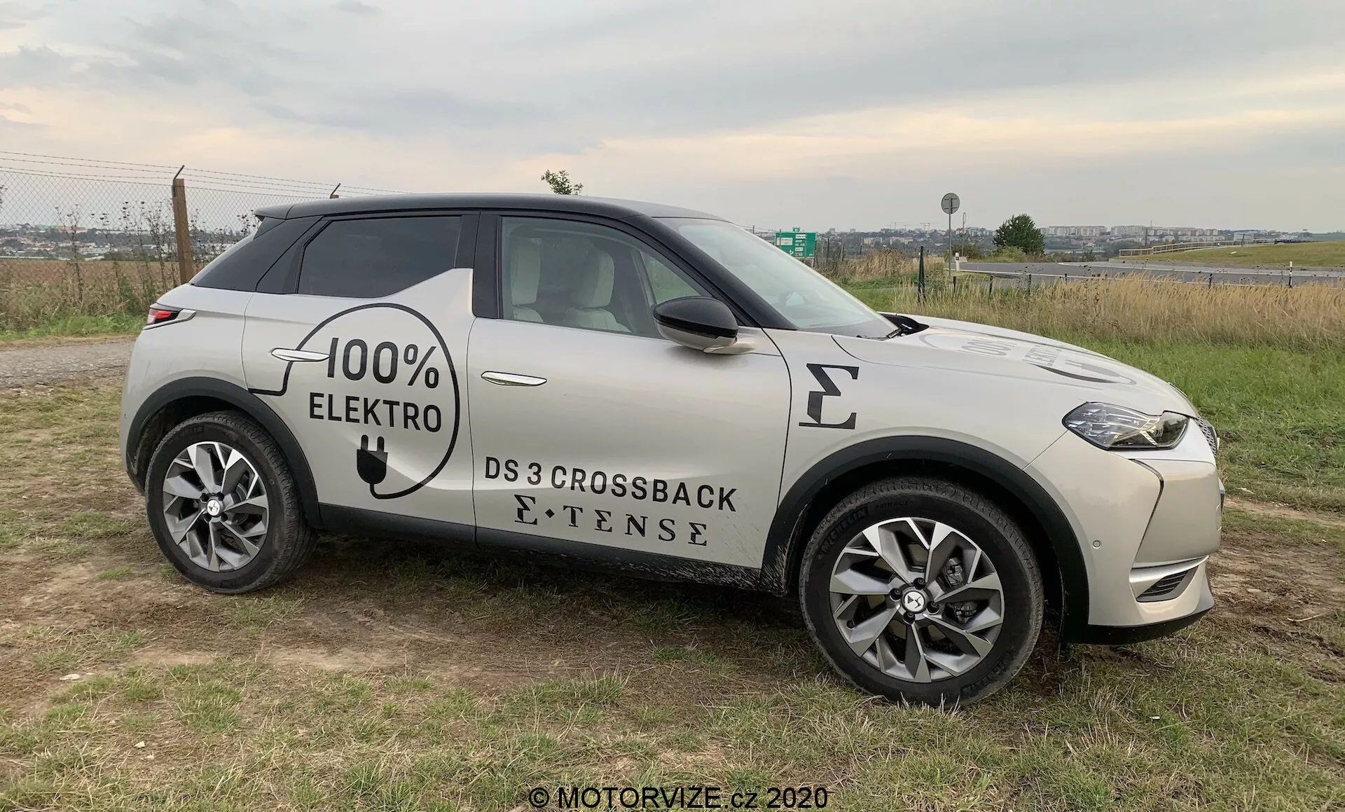 Stranski profil sodobnega SUV-ja, DS 3 Crossback E-Tense iz leta 2019, električnega vozila z grafiko '100% ELEKTRO' na boku. Vozilo je nameščeno na travnati površini z nekoliko asfaltne ceste v ozadju, kar omogoča jasen pogled na prednjo mrežo, LED žaromete, sprednje kolo in profil pod lahkim kotom. Vidne funkcije vključujejo večkraka lita platišča, stranska ogledala z vgrajenimi indikatorji, vdolbene kljuke vrat in simbol polnilnega mesta na sprednjem blatniku, kar nakazuje poudarek na napredni tehnologiji, slogu in ekološki osredotočenosti, vse pod naravno, oblačno svetlobo.