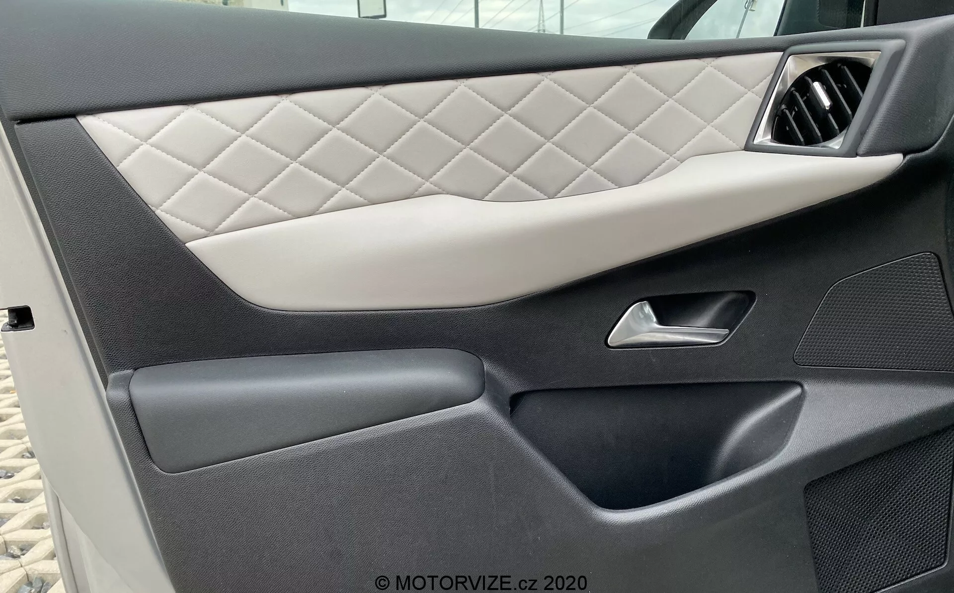 Pogled od blizu na notranjo ploščo vrat vozila DS Automobiles DS-3 2019 (DS-3-Crossback-E-Tense), ki prikazuje prešito oblazinjenje z diamantnim vzorcem, elegantno srebrno kljuko, temne visokokakovostne plastične dele z različnimi teksturami in spodnji brezšivni žep za shranjevanje, poudarjajoč luksuzen in podroben dizajn avtomobila.