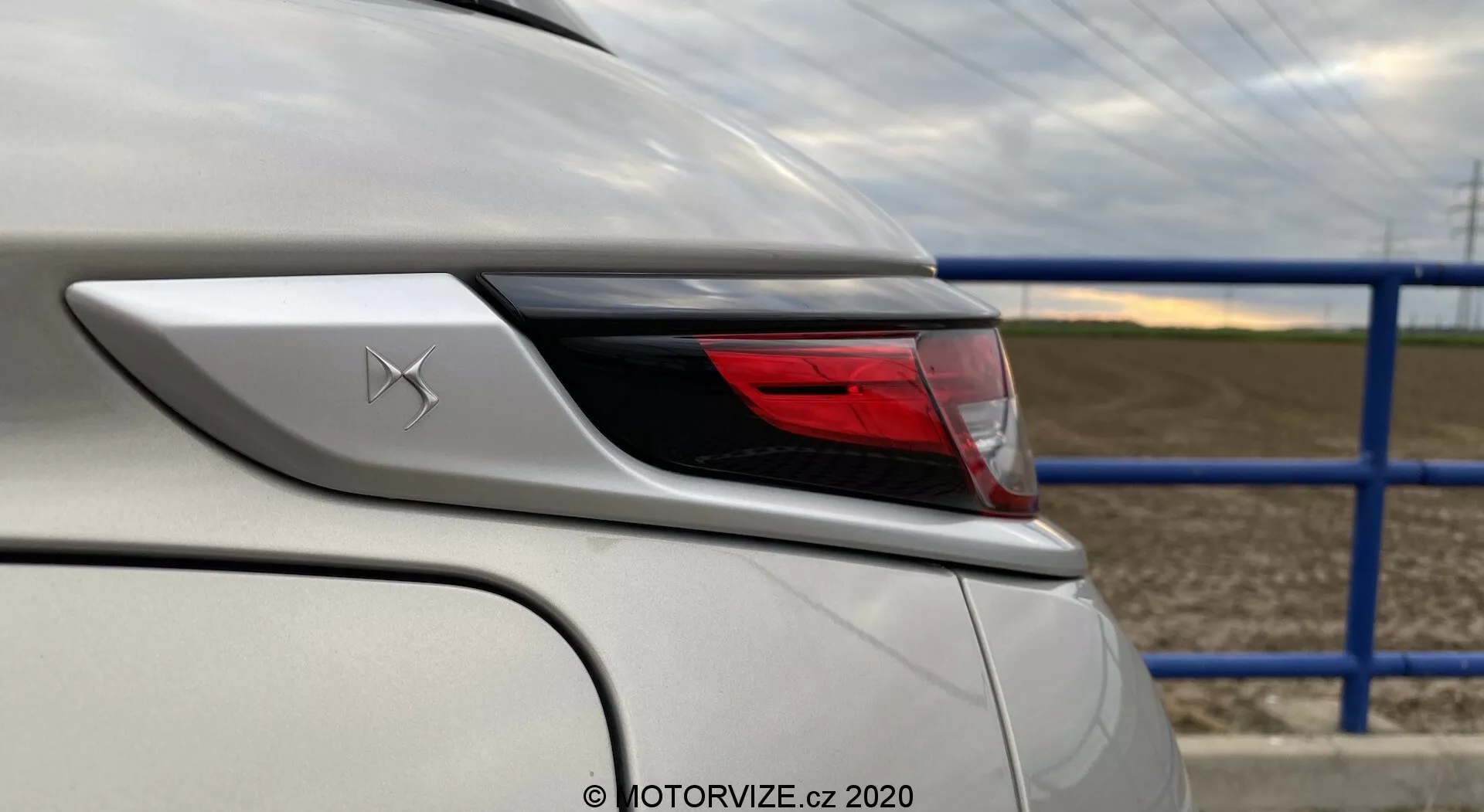 Podroben pogled na zadnji kot belega vozila DS-3 2019 (DS-3 Crossback E-Tense), osredotočen na oblikovne elemente in funkcije okoli zadnje svetlobne skupine. Fotografija iz zadnjega četrtnega kota z leve strani poudarja ukrivljenost zadnje svetlobne skupine in zadnjega brizgalnika, prikazuje podrobnosti karoserije vozila in napredno LED osvetlitveno sestavo. Nad zadnjo svetlobno skupino je viden emblem s stiliziranim X, bela barva vozila je brezhibna in odseva sijajni zaključek. V ozadju je cesta z modro kovinsko ograjo in kmetijsko polje.