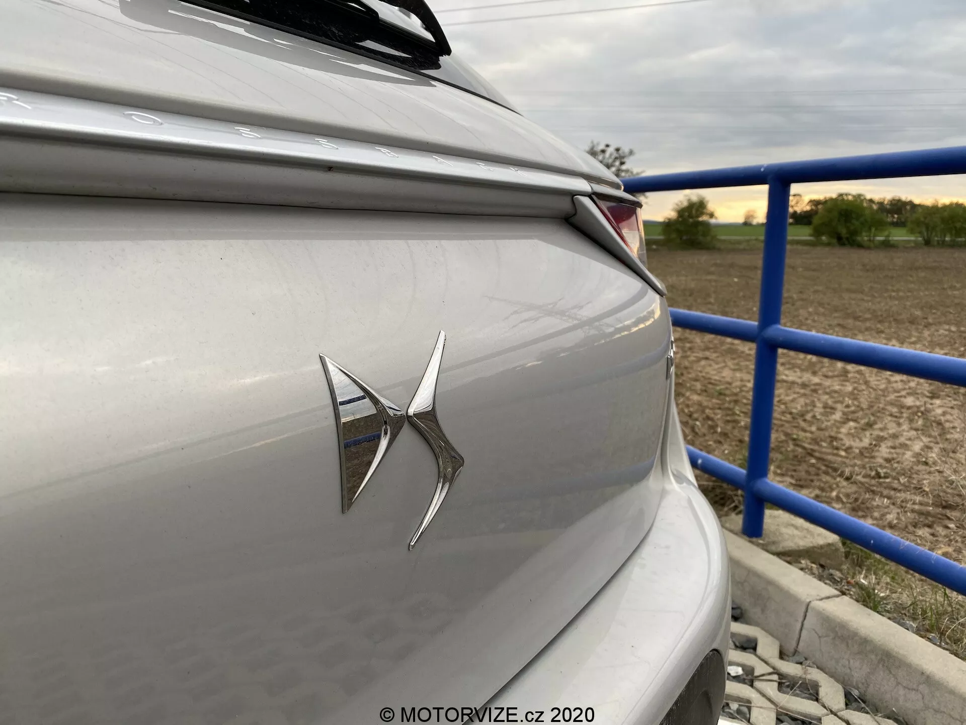 Bližnji posnetek zadnjega dela luksuzne limuzine ali hatchbacka svetlo srebrne ali sive barve, z vidnim emblemom blagovne znamke na prtljažniku in modernim LED zadnjim svetilnim sklopom. Fotografija, posneta z rahlim kotom blizu zadnjega desnega dela vozila, prikazuje ostre horizontalne oblikovalske linije in predmestno ali podeželsko ozadje z modrimi ograjami in strukturami. Oblikovalna estetika avtomobila poudarja sodobno tehnologijo in nevtralno, umirjeno barvno shemo.