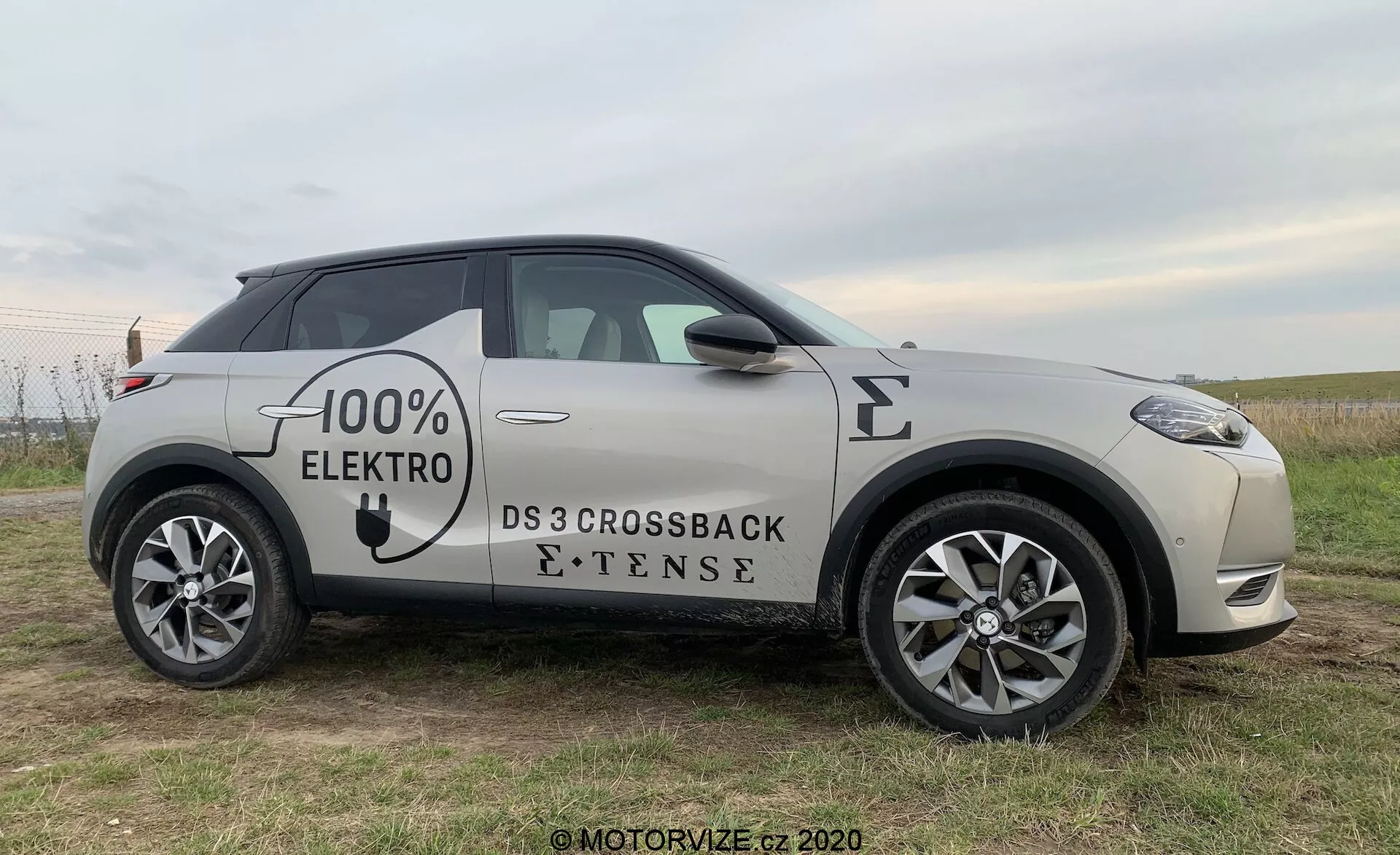 Stranski profil električnega vozila DS Automobiles DS 3 Crossback E-Tense 2019, fotografiran iz rahlega sprednjega kota, ki poudarja sprednje kolo, voznikova vrata in ogledalo, z delnimi pogledi na sprednjo masko in zadnje kolo, postavljen na travnato polje z subtilnim ozadjem.
