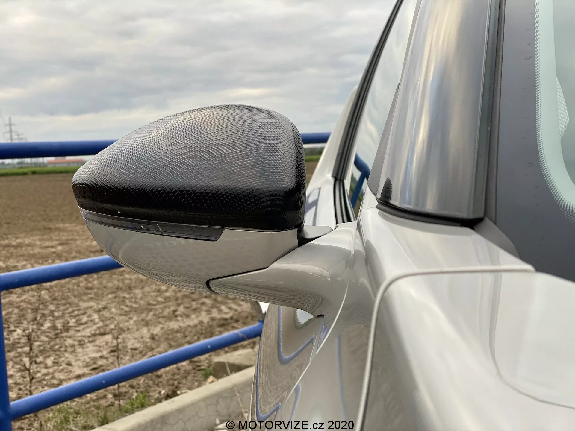 Pogled od blizu na zunanje ogledalo voznikovih vrat avtomobila ds-automobiles ds-3 2019 (ds-3-crossback-e-tense). Ogledalo je pobarvano tako, da ustreza barvi karoserije, razen zgornjega dela ohišja, ki ima vzorec iz karbonskih vlaken, kar kaže na premium model. Pod ogledalom je delno viden vratni panel v barvi karoserije in vidna je kontura. Ogledalo vključuje sodobne oblikovne elemente, vključno z vgrajeno LED smerno lučko. Slika se osredotoča na podrobnosti oblikovanja ogledala z zamegljenim ozadjem, ki prikazuje odprto polje in kovinsko pregrado, kar kaže na podeželsko okolje.