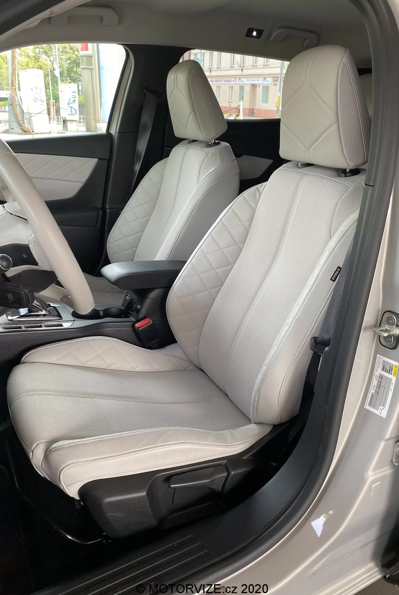 Notranjost DS Automobiles DS 3 Crossback E-Tense 2019, osredotoča se na voznikov sedež iz zunanje perspektive na strani voznika. Slika, posneta iz rahlo nizkega do visokega kota, prikazuje sodoben dvoobarvni usnjeni sedež s svetlo sivo sredino in temno sivimi ali črnimi robovi. Sedež je očitno zasnovan za udobje in športno estetiko, ima izrazite stranske ojačitve, vidne šive in elektronske nastavitvene kontrole. Odprta voznikova vrata, vidni varnostni pas, delno vidna sredinska konzola in vratna plošča z različnimi kontrolnimi gumbi kažejo na premium materiale in številne napredne funkcije. Dnevna osvetlitev izboljšuje jasnost luksuznih notranjih podrobnosti.