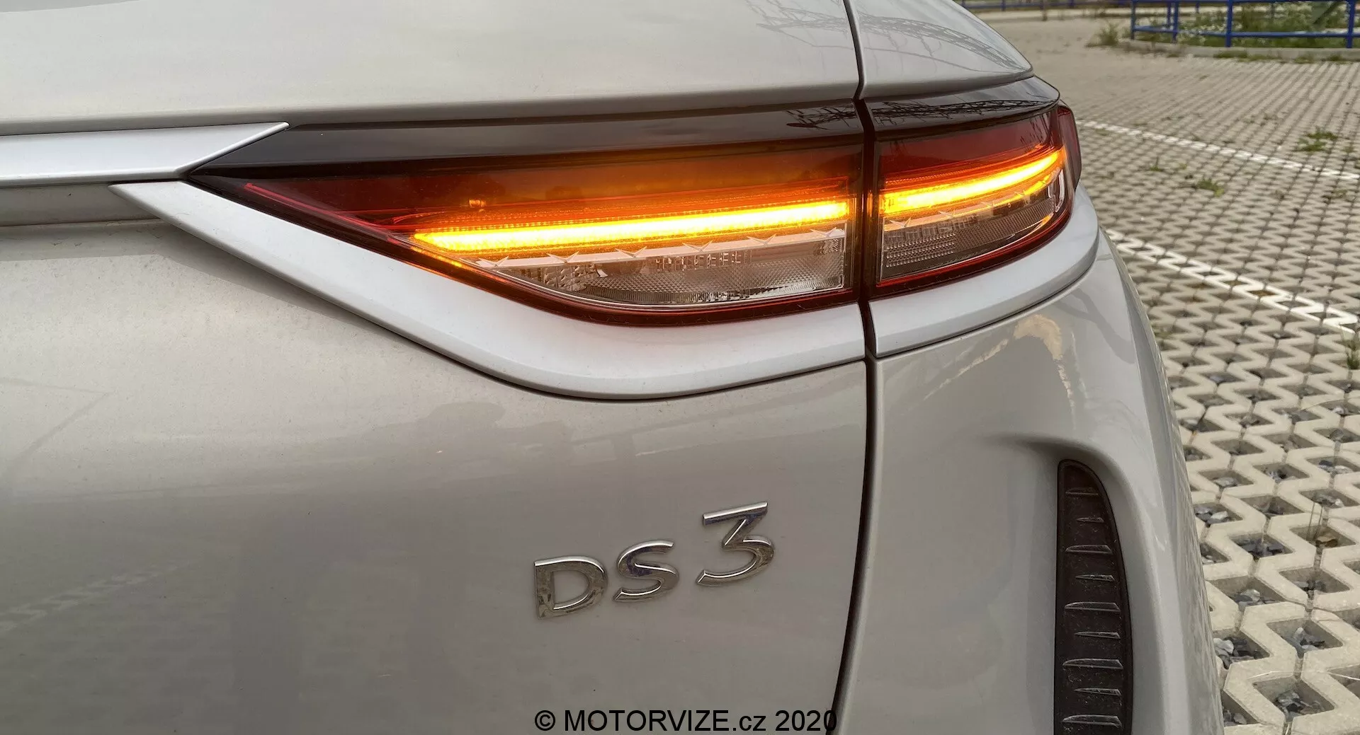 Pogled od blizu na zadnji del DS 3 Crossback E-Tense (model 2019), ki prikazuje zadnjo desno četrtino iz rahlo dvignjenega kota. Slika poudarja LED zadnjo luč in smernik z živo oranžno osvetlitvijo. Vidni elementi vključujejo zgornji rob prtljažnika, ukrivljenost zadnjega blatnika, natančno poravnavo plošč karoserije ter del desnega zadnjega kolesa. Avtomobil ima srebrno ali svetlo sivo barvo. Fotografija posneta na teksturirani površini.