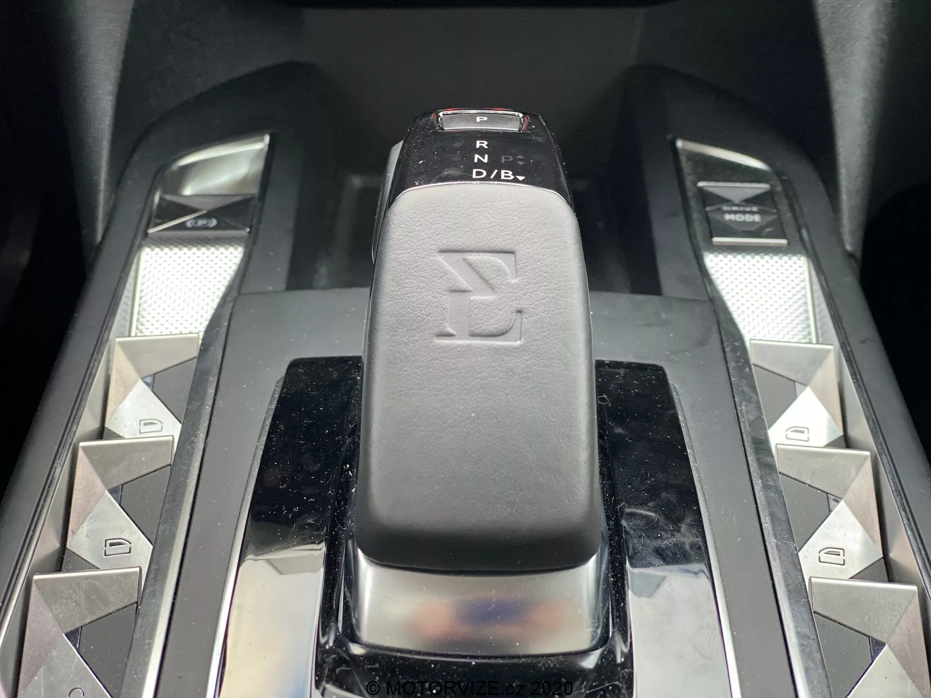 Podroben posnetek prestavne ročice v DS Automobiles DS-3 2019 (DS-3 Crossback E-Tense), ujet z rahlo dvignjenega sprednjega kota. Prestavna ročica, z elegantno črno usnjeno ali usnju podobno površino z indikatorji PRND, je v središču slike. Simetrični srebrni gumbi v obliki diamantov in dva kovinska drsnika obkrožata ročko in nakazujeta dodatne funkcije avtomobila. Sijoči in vrhunski materiali izžarevajo sodobno, razkošno notranjost avtomobila.