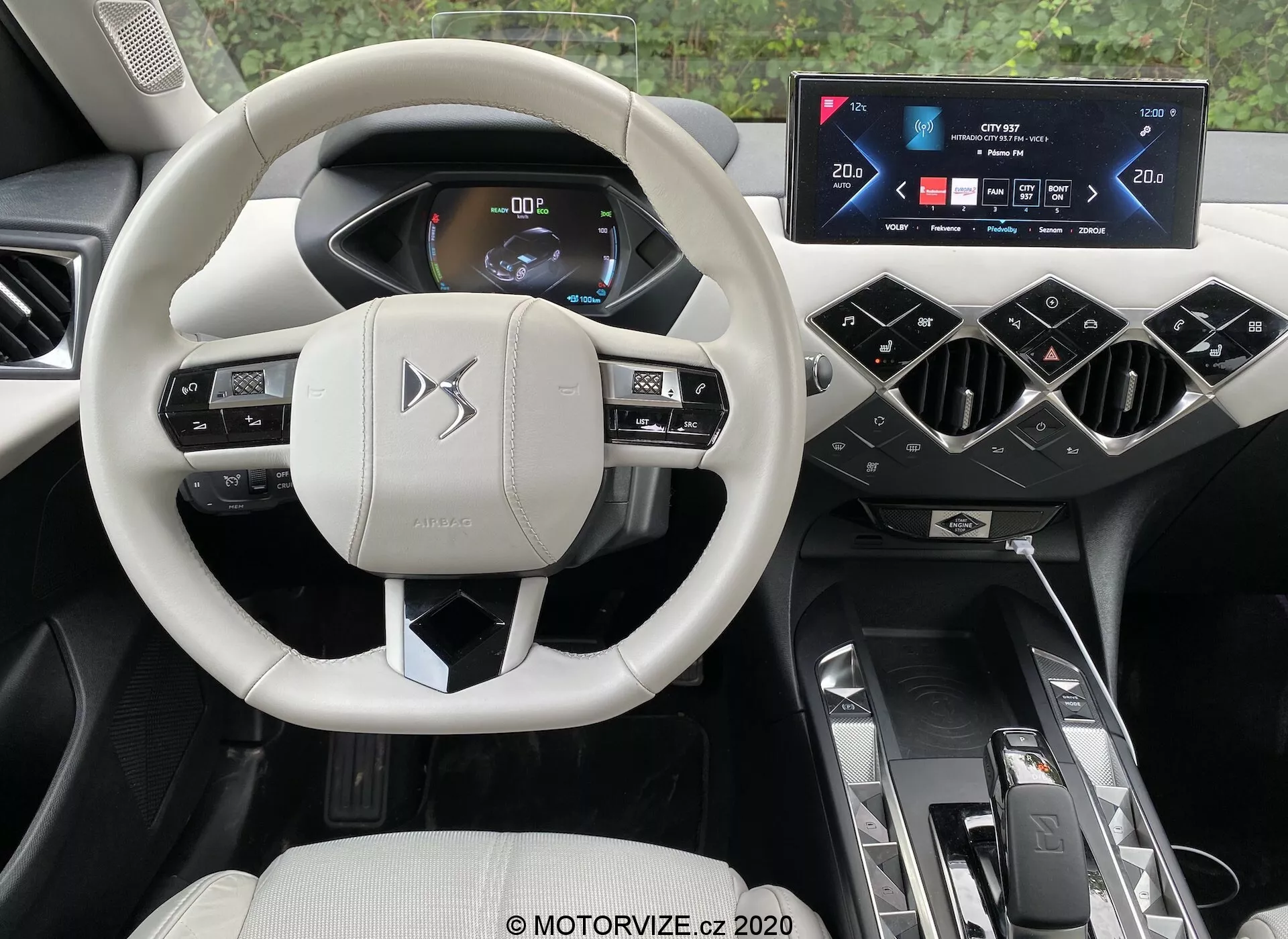 Pogled v notranjost modernega avtomobila iz voznikove perspektive, poudarjajoč sofisticirane elemente ds-automobiles ds-3 2019 (ds-3-crossback-e-tense). Osrednji del je svetlo obarvan, večfunkcijski volan z nadzornimi gumbi in lopatkami, postavljen proti ozadju digitalnega instrumentnega sklopa, ki prikazuje informacije o vozilu z jasno, svetlo grafiko. Nad sredinsko konzolo se nahaja širokozaslonski zaslon na dotik za informacijsko-zabavni sistem, obdan s poševnimi prezračevalnimi režami. Sredinski predor vključuje samodejnega izbirnika prestav, nadzor načinov vožnje in dvojne nosilce za pijače. Razkošna notranjost vrhunske kakovosti vključuje svetle in temne tone, ki ponazarjajo udobje, priročnost in napredno avtomobilsko tehnologijo.