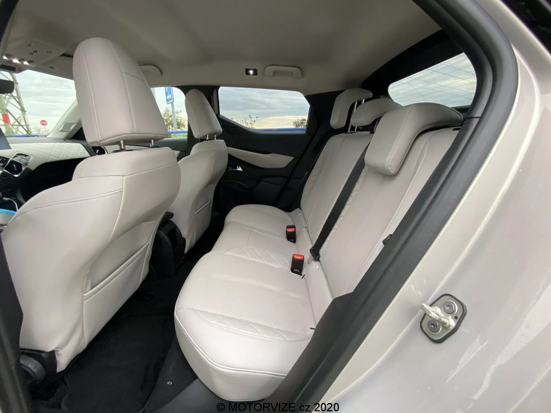 Notranji pogled DS Automobiles DS 3 Crossback E-Tense 2019 iz poševnega kota, osredotočen na zadnji potniški prostor. Svetle usnjene sedeže, vidne vzglavnike, varnostne pasove in sredinski zložljiv naslon za roke na zadnjem sedežu nakazujejo luksuzno in dobro vzdrževano notranjost. Temne preproge kontrastirajo s svetlim oblazinjenjem, električni upravljalnik oken pa je viden na vratih. Kot kamere rahlo navzdol iz zadnjih desnih vrat poudarja sedežno površino in prostor za noge.