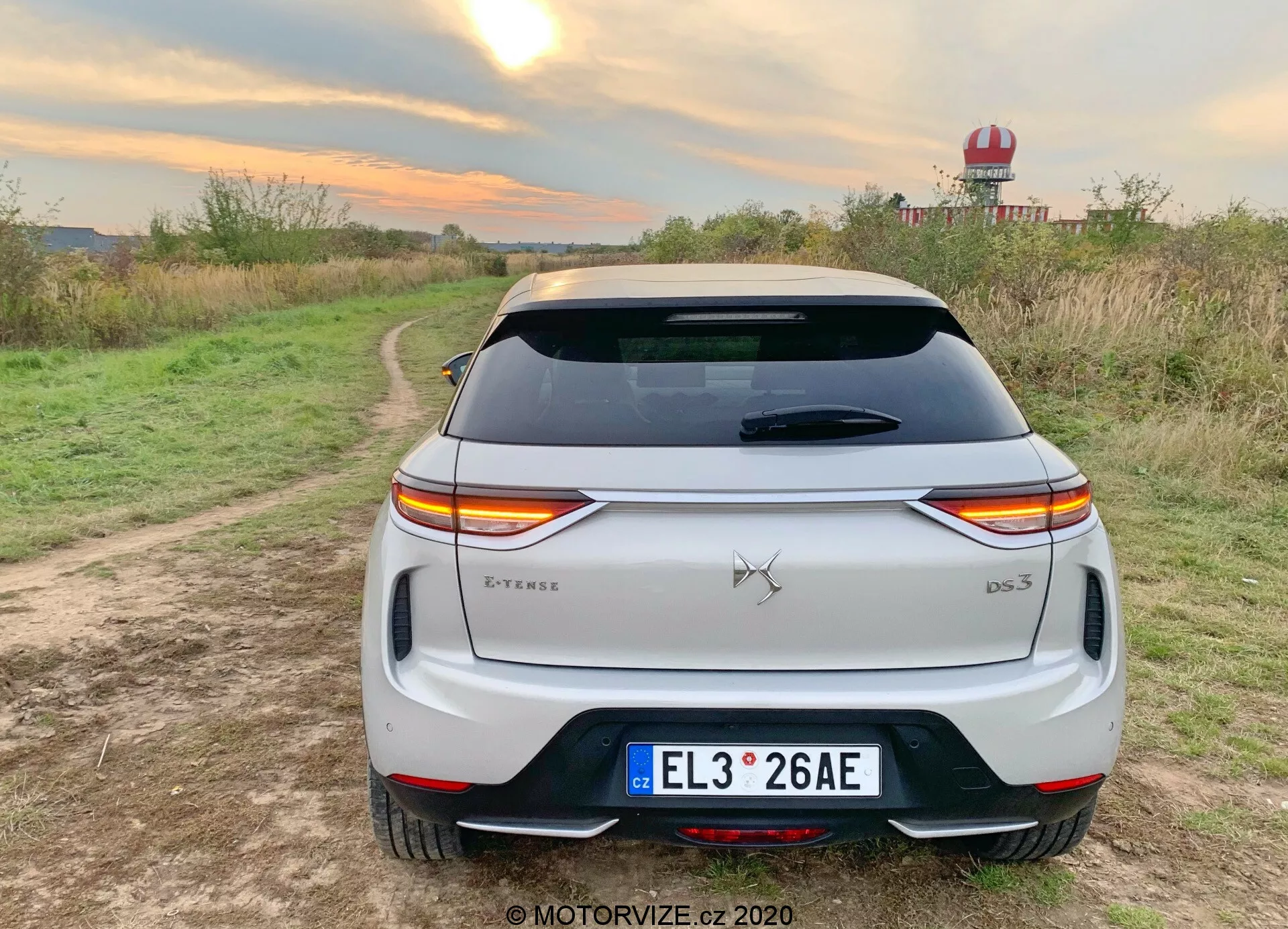 Pogled od zadaj na bel 2019 DS Automobiles DS 3 Crossback E-Tense crossover SUV parkiran na makadamski poti z rastlinjem na obeh straneh v podeželskem okolju. Značilne LED zadnje luči se raztezajo čez celoten zadnji del vozila, prekinjene z emblemom znamke. Oznaka modela 'DS 3' je nameščena v spodnjem desnem kotu prtljažnika, medtem ko je oznaka 'E-Tense' na spodnjem levem delu, kar nakazuje električni ali hibridni pogonski sklop. Lastnosti vključujejo evropsko registrsko tablico, zadnje steklo z linijami za odstranjevanje mrzlišča, strešni spojler, zadnji odbijač z vgrajenimi odbojniki in črno obrobo ter dvojne izpušne cevi, kar kaže na bolj športni dizajn. Okolje nakazuje na testno vožnjo ali življenjski stil fotografiranju, ki poudarja uporabo na prostem, s toplimi svetlobnimi pogoji, ki poudarjajo belo barvo vozila.