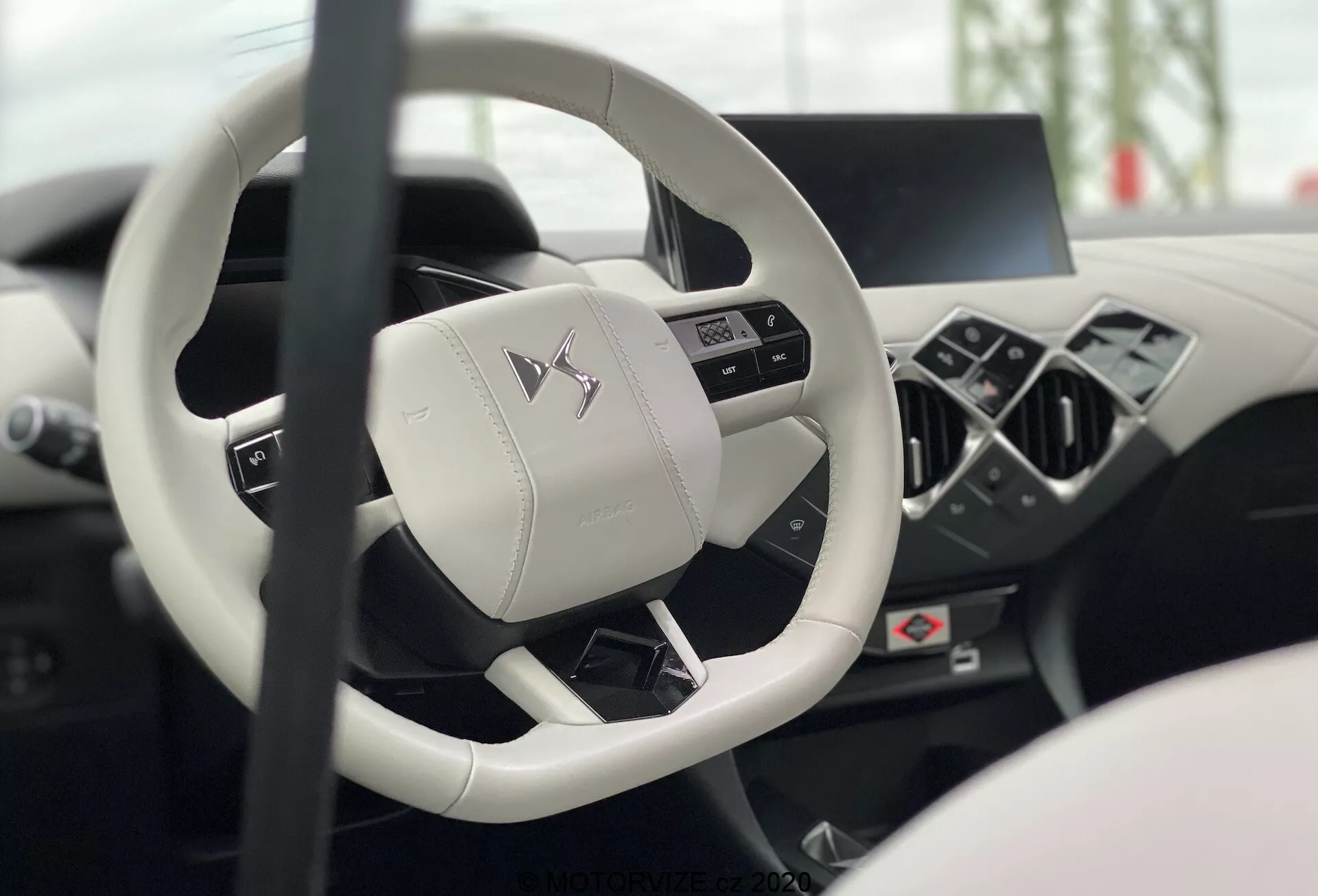 Notranjost DS Automobiles DS-3 2019 (DS-3-Crossback-E-Tense) iz voznikovega sedeža, s poudarkom na sodobnem volanu s tremi kraki in večfunkcijskimi gumbi ter zamegljeno digitalno armaturno ploščo in sredinsko konzolo, ki kažeta luksuz in napredno tehnologijo