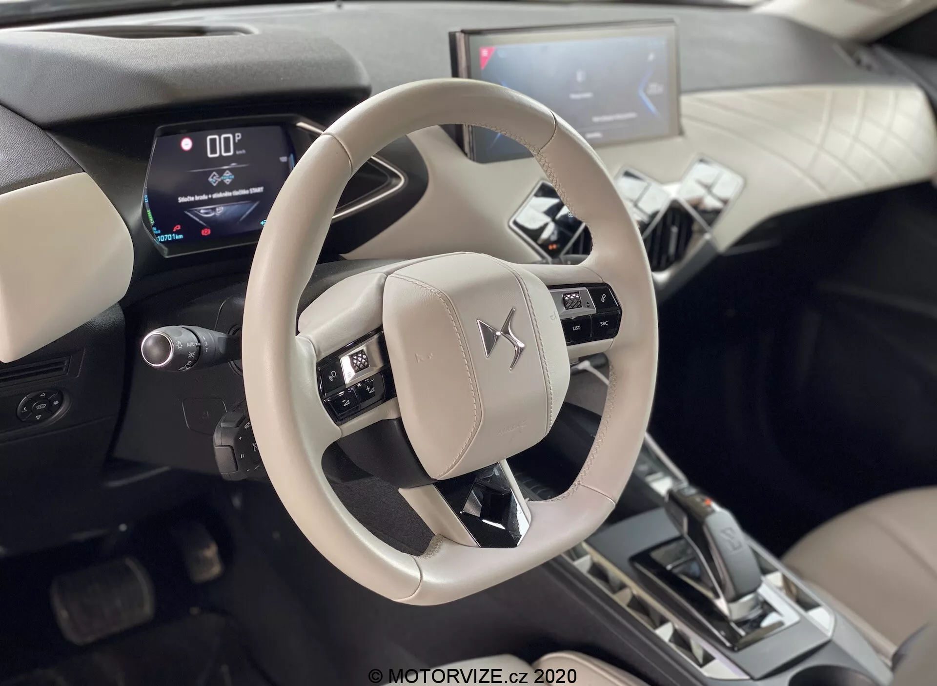 Notranji posnetek DS-Automobiles DS-3 2019 (DS-3 Crossback E-Tense) osredotočen na svetlo bež/sivo volan z Mitsubishi emblemom in osrednjo konzolo. Posneto z vidika zadnjih sedežev, prikazuje nameščene kontrole za avdio, glasovno in tempomat sisteme. Digitalno instrumentno ploščo prikazuje hitrost nič in prestavno ročico v položaju 'P', kar nakazuje, da je vozilo parkirano. Osrednji infotainment sistem z vidnim zaslonom na dotik v dvobarvni notranjosti z kovinskimi poudarki, kar kaže na sodobne digitalne prikazovalnike in napredne avtomobilske funkcije.