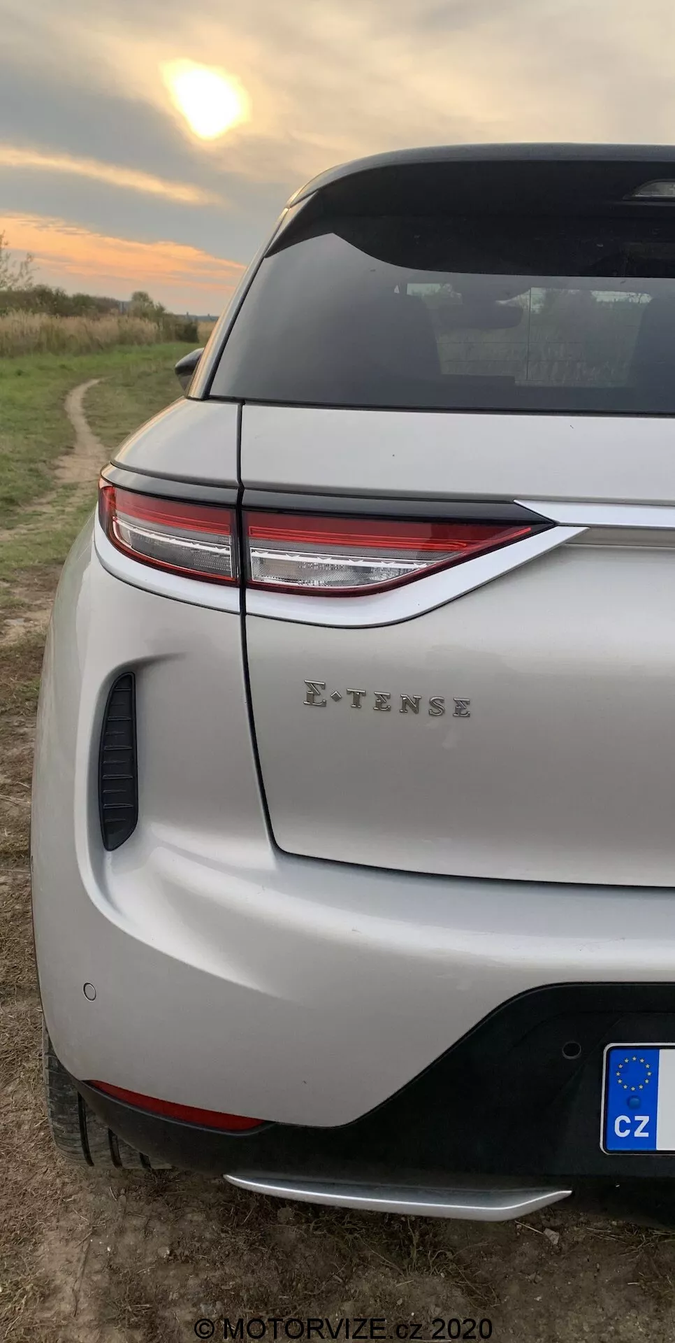 Vertikalni bližnji posnetek zadnjega vogalnega dela avtomobila DS-3 2019 (DS-3 Crossback E-TENSE). Osredotoča se na sodobne LED zadnje luči, športni zadnji odbijač z integriranim difuzorjem in parkirnim senzorjem, pokrov prtljažnika z aerodinamičnim spojlerjem in delni pogled na zadnje kolo in blatnik. Nameščeno je na podeželskem ozadju z odprtim poljem ob sončnem vzhodu ali zahodu, kar kaže na sodobno zasnovo vozila in njegove električne zmožnosti.