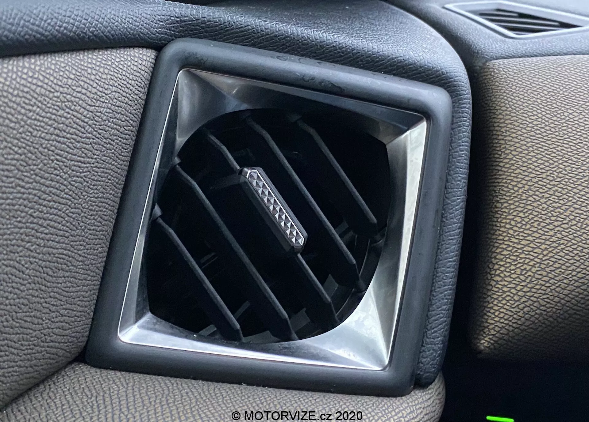 Pogled od blizu na zračno odprtino v notranjosti vozila DS Automobiles DS-3 Crossback 2019, ki prikazuje nastavljive vodoravne letvice znotraj diamantne odprtine, obdane s teksturirano plastično površino armaturne plošče s kvadratnim robom za dodatno oblikovanje.