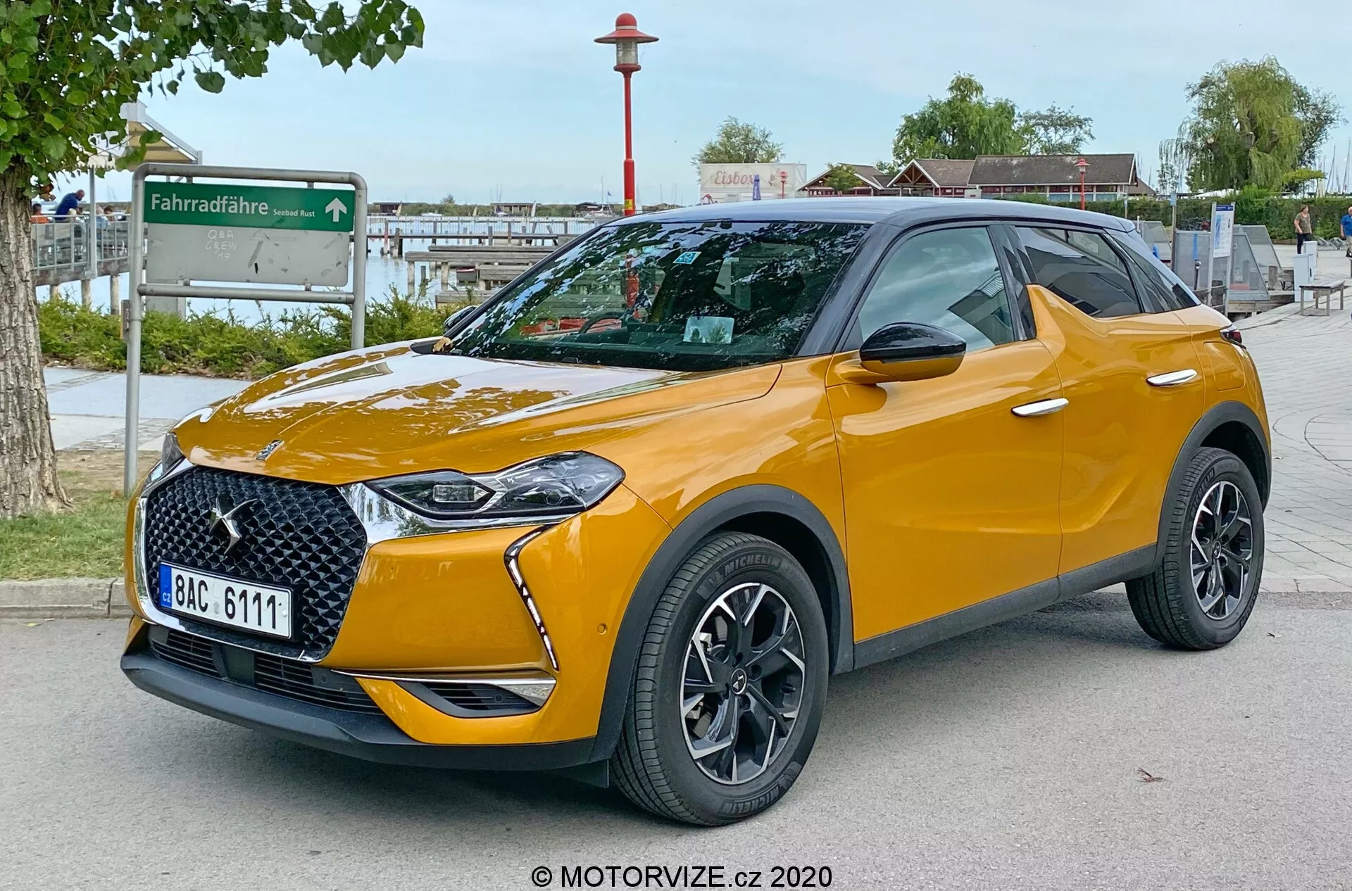 Slika prikazuje sodoben SUV DS Automobiles DS 3 Crossback 2019 v pogled s treh četrtin spredaj, ki poudarja elegantno prednje rešetko, integrirane LED žaromete, sprednji odbijač, pokrov motorja in profil voznikove strani. Parkiran na prostem na tlakovanem površju z urbanim ozadjem, ki vključuje postajo za deljenje koles. Fotografija, posneta z nizkega zornega kota na višini žarometov, poudarja oddaljenost avtomobila od tal. Vidne lastnosti vključujejo večkrake aluminijeste platišča, integrirane smerne luči v ogledalih, nizkoprofilne pnevmatike, strešni kontrast z nosilci za streho, kovinski zaključek barve, značke specifične za trim in zatemnjena zadnja okna. Registrska tablica je namerno zamegljena za zasebnost.
