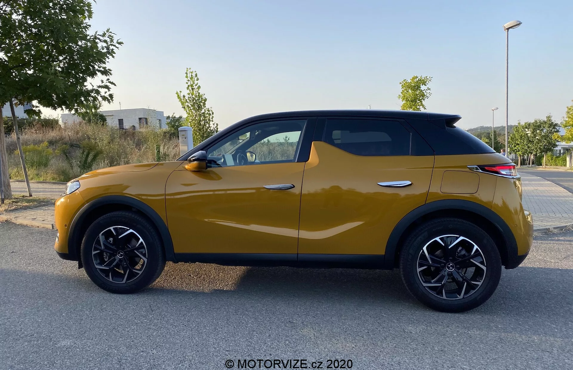 Stranski profil vozila DS Automobiles DS 3 Crossback iz leta 2019 v gorčično rumeni barvi. Moderni kompaktni SUV predstavlja značilne črne obrobe kolesnih lokov, večkrake črne aluminijaste platišča in dinamično linijo karoserije, ki se razteza od prednjega kolesnega loka do zadnjih luči. Vzvratna ogledala imajo vgrajene smerne kazalnike, okna obrobljena s črno obrobo pa poudarjajo usklajen dizajn, medtem ko naklonjena streha dodaja sodoben pridih. Slika, posneta pri difuzni svetlobi, ne prikazuje senc ali bleščanja, kar omogoča osredotočenost na oblikovne elemente vozila.