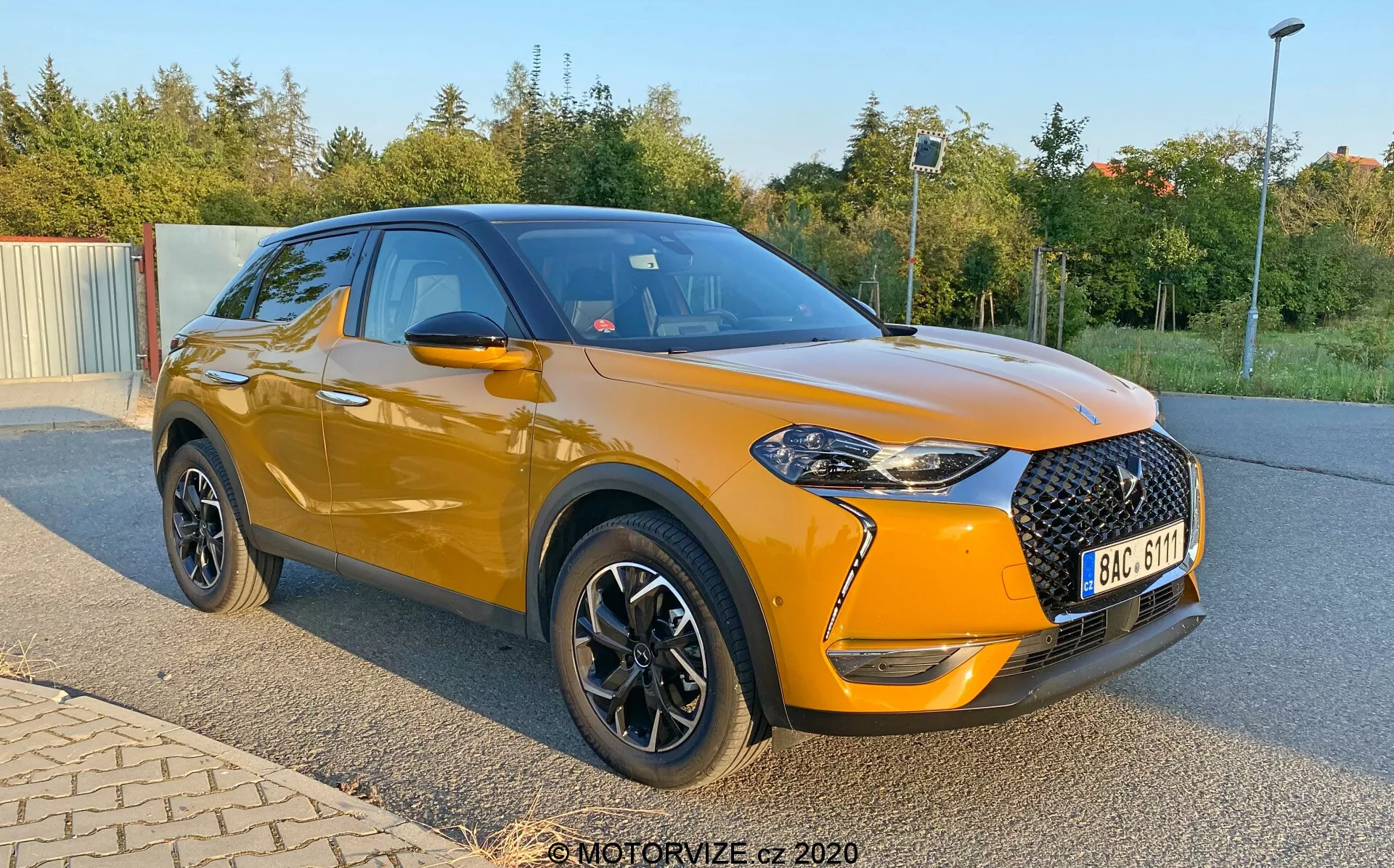 Moderen, kompakten SUV DS Automobiles DS-3 Crossback iz leta 2019, prikazan parkiran pod kotom s poudarkom na sprednjem in stranskem profilu. Slika, posneta iz nižje perspektive blizu kolesnih pest, poudarja svetlo oranžno kovinsko ohišje vozila in črno streho. Sprednji del ima veliko črno mrežo z diamantnim vzorcem, ostre žaromete z LED poudarki in meglenke v črnih okvirjih. Avto ima črna stranska ogledala, kromirane okenske obrobe in črna kolesa s srebrnimi poudarki. Opazna je rahlo dvignjena drža, športne linije karoserije in guba, ki se spušča čez kljuke vrat.