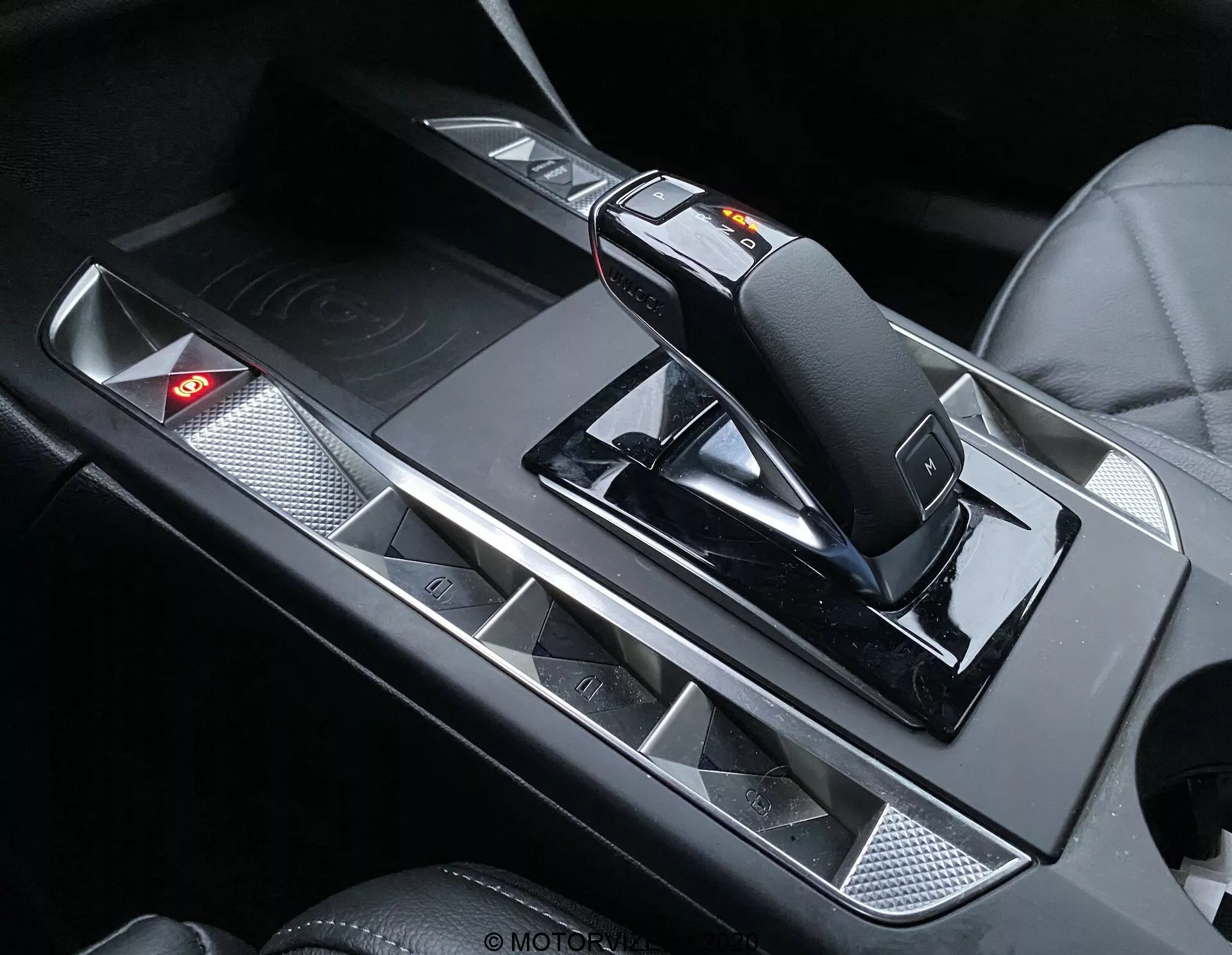 Pogled od blizu na notranjost DS Automobiles DS-3 Crossback 2019, osredotočen na osrednjo konzolo z moderno, ergonomsko prestavno ročico s stikalom za ročno prestavljanje in sprostitvenim mehanizmom. Konzola ima kovinske poudarke, ki kontrirajo s črnimi površinami, držala za pijačo z mat površino in nadzorno ploščo z osvetljenim rdečim opozorilnim lučom.