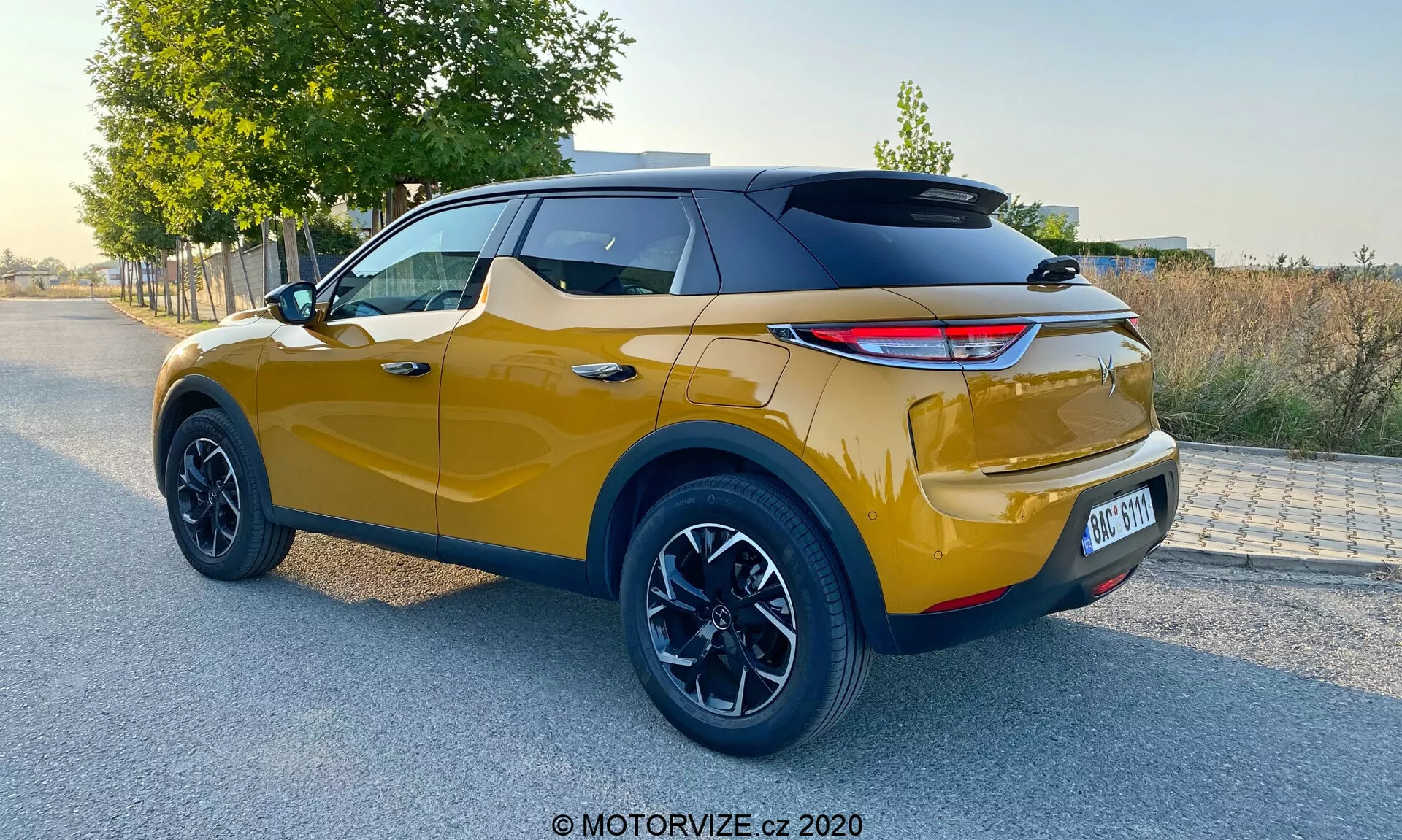 Zadnji tričetrtinski pogled na sodoben crossover SUV v naravni dnevni svetlobi, z bleščečo kovinsko zlato barvo in športno črno streho, modernimi LED zadnjimi lučmi, velikimi črnimi aluminijastimi platišči, visoko oddaljenostjo od tal, vidnimi zadnjimi parkirnimi senzorji in dvobarvnim dizajnom, značilnim za model DS Automobiles DS 3 Crossback leta 2019.