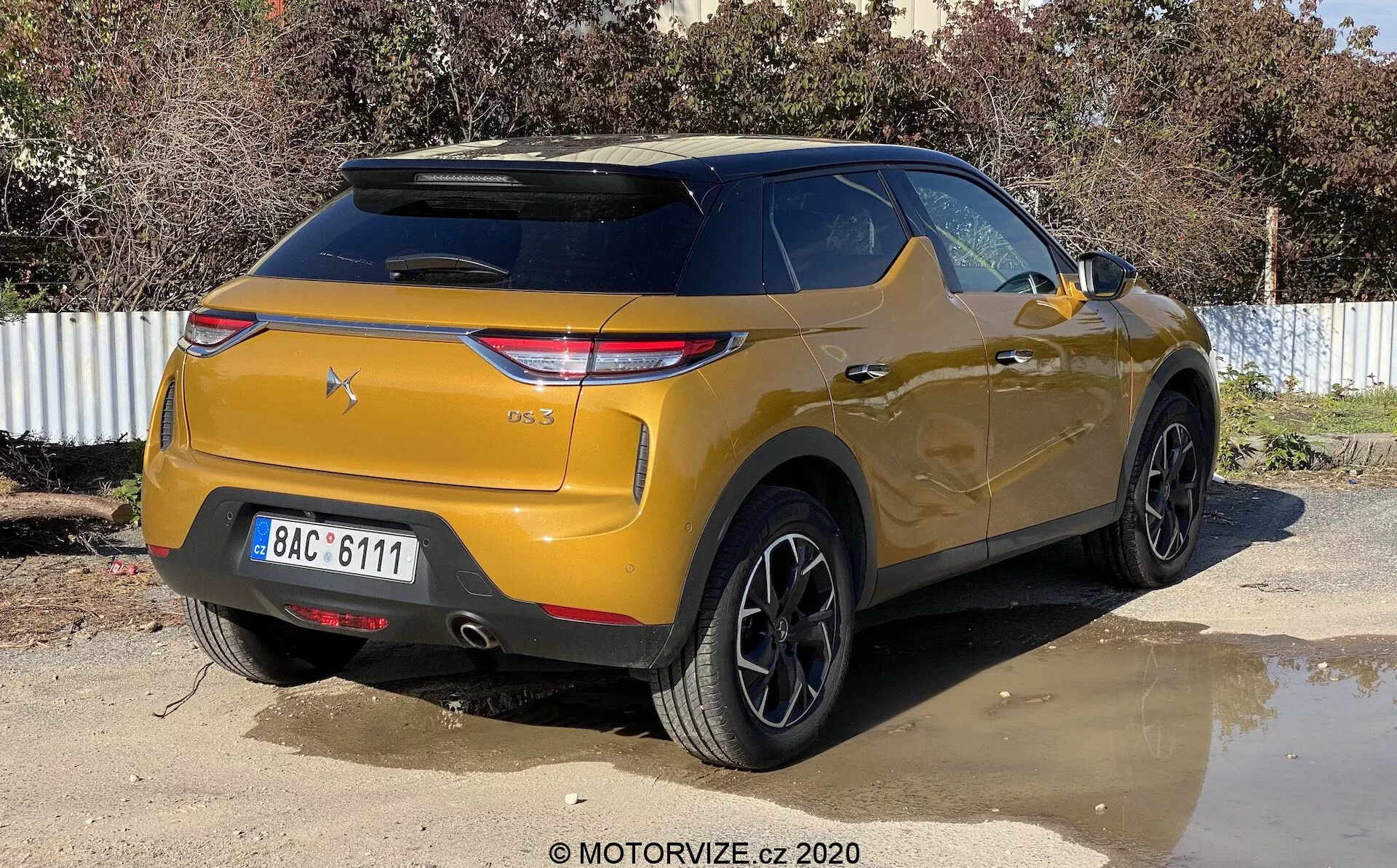 Zadnji trije četrt pogled na kovinsko zlate barve DS-3 Crossback SUV, parkiran na prostem na sončen dan. Slika zajame vozilo z rahlim desnim kotom, poudarja desno zadnjo stran in prtljažno območje. Ključne lastnosti vključujejo moderne kotne zadnje svetlobne sklope, nagnjeno zadnje vetrobransko steklo z integriranim strešnim spojlerjem, izrazit odbijač s parkirnimi senzorji, črna več-kraka lita platišča, visoko oddaljenost od tal in zatemnjena zadnja okna.