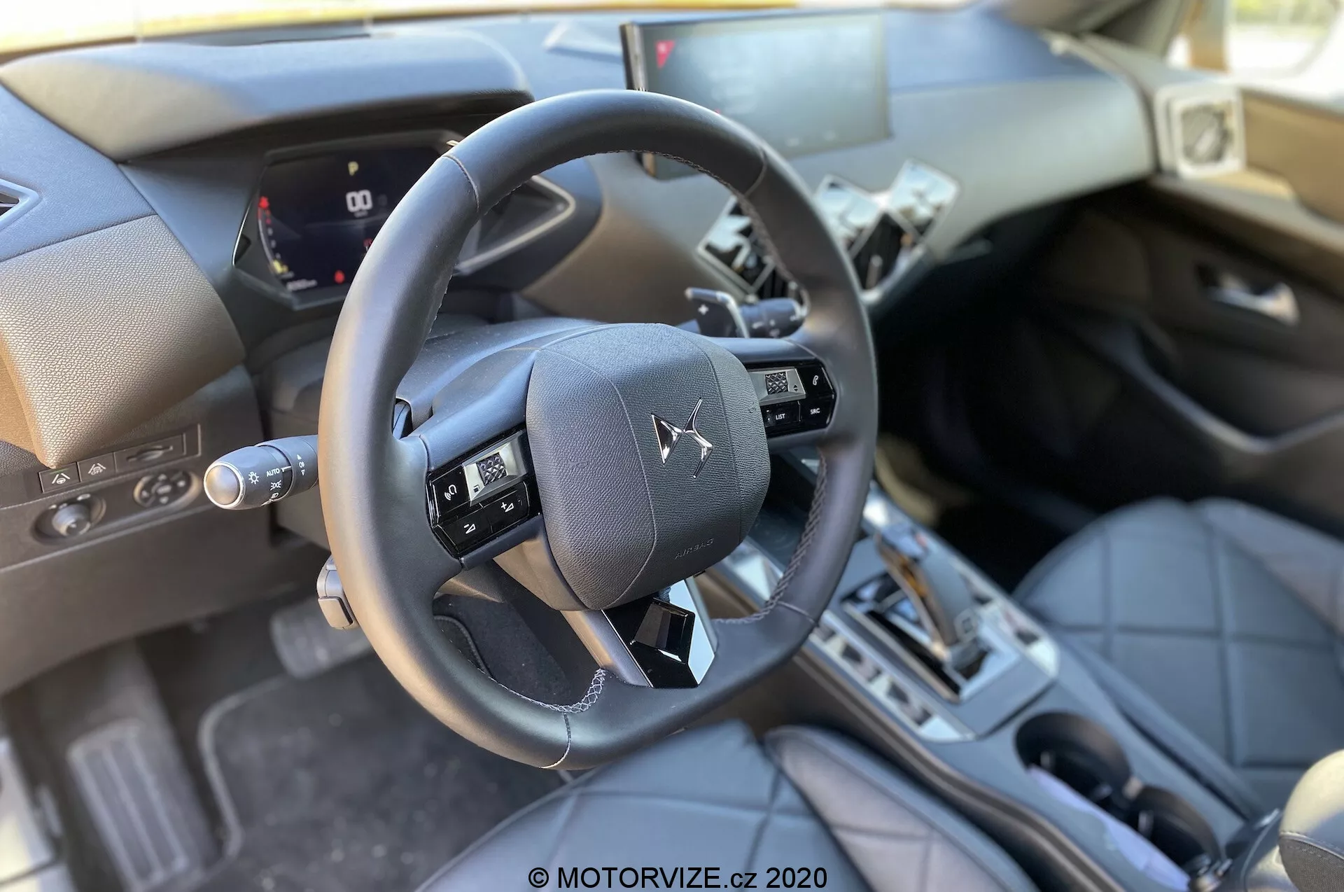 Notranji pogled DS Automobiles DS 3 Crossback 2019 iz perspektive sprednjega sedeža sovoznika, poudarjajoč trikraki volan z večfunkcijskimi gumbi, delni digitalni instrumentni grozd, ki prikazuje hitrost avtomobila in merilnike, sredinsko konzolo z avtomatskim menjalnikom, vrhunske materiale, kot so usnjene površine in kovinski poudarki, tradicionalno režo za vžig, pedale in dobro osvetljeno naravno dnevno svetlobo.