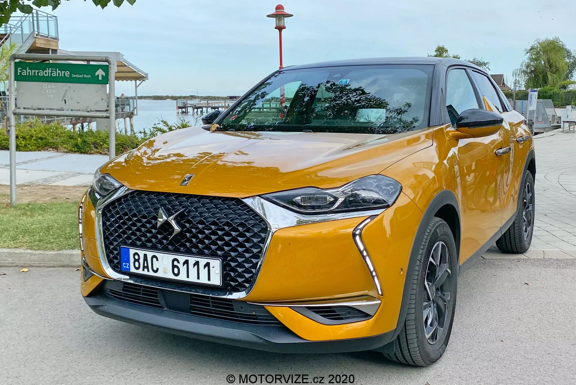 Slika prikazuje DS Automobiles DS 3 Crossback 2019, moderen kompakten crossover SUV, parkiran na tlakovanem obcestnem območju. Posneta iz nizkega kota spredaj od strani, slika poudarja sprednji del vozila in njegovo desno stran. Ključne značilnosti vključujejo izrazit emblem blagovne znamke na mreži z diamantnim vzorcem, elegantne LED žaromete s stiliziranimi dnevnimi lučmi, integrirane meglenke na prednjem odbijaču, ogledala v barvi karoserije s smerniki in temna večkraka lita platišča. Avto je prebarvan v živo kovinsko oranžno barvo s poudarjenimi linijami karoserije, ki izboljšujejo njegov aerodinamični videz, ter oznake, ki morda navajajo stopnje opreme. Okolica nakazuje javno območje v bližini parka, kar dokazuje znak kolesarske poti v ozadju.