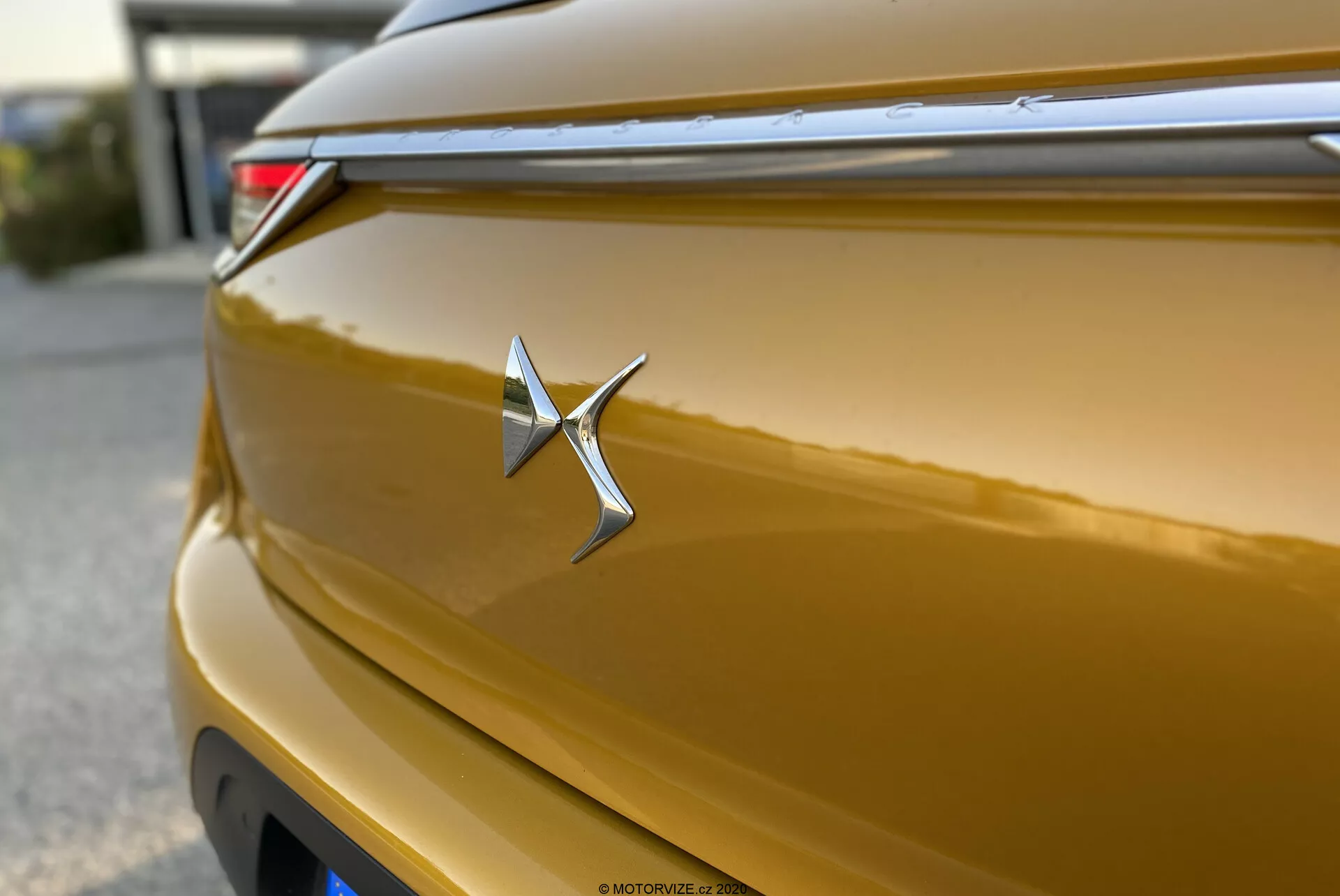 Posnetek od blizu značke DS Automobiles DS-3 Crossback 2019 v kovinsko zlati barvi. Kromiran emblem je oster in osrednje postavljen, spominja na zvezdo z podaljšanim desnim krakom. Sijajna karoserija odseva svetlobo po svojih krivinah, prikazuje bogat zlat odtenek. Plitka globinska ostrina poudarja značko, s horizontalno črto nad njo, ki prispeva k aerodinamičnemu dizajnu. Del zadnje LED luči je delno viden v zgornjem desnem kotu. Zadnji pokrov ima subtilno ukrivljenost, efekt bokeh v ozadju pa izolira značko.