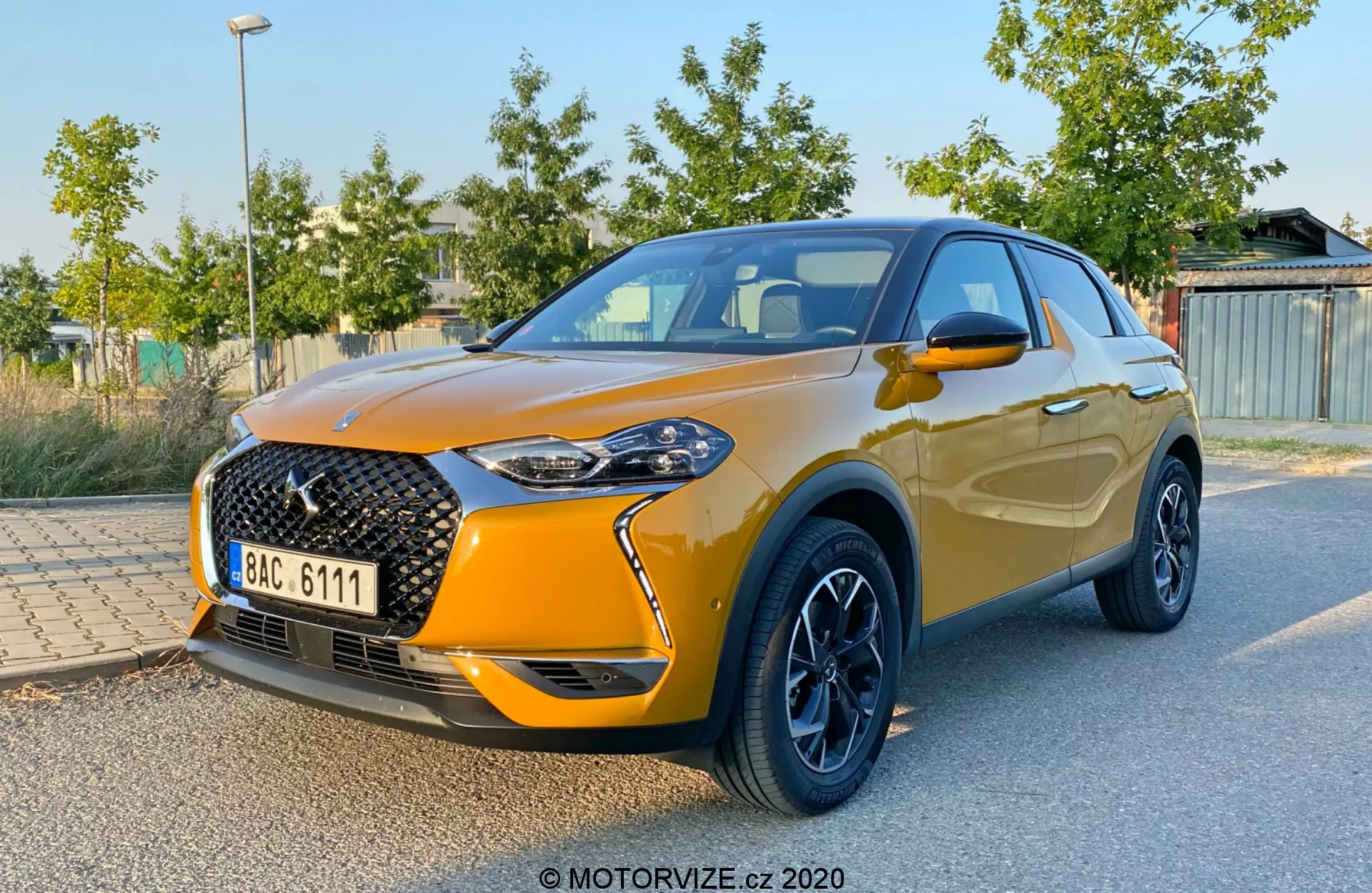 Sprednji pogled na DS Automobiles DS 3 Crossback iz leta 2019, ki prikazuje značilno veliko sprednjo mrežo z mrežastim vzorcem in izrazitim znakom, elegantne LED žaromete, kovinsko oranžno barvo, kontrastna črna lita platišča, integrirana stranska ogledala in vzpenjajočo se linijo oken, ki se konča z aerodinamičnim spojlerjem. Vozilo je parkirano z zelenim listjem in strukturami v zamegljenem ozadju, kar poudarja dizajn in športne lastnosti avtomobila.