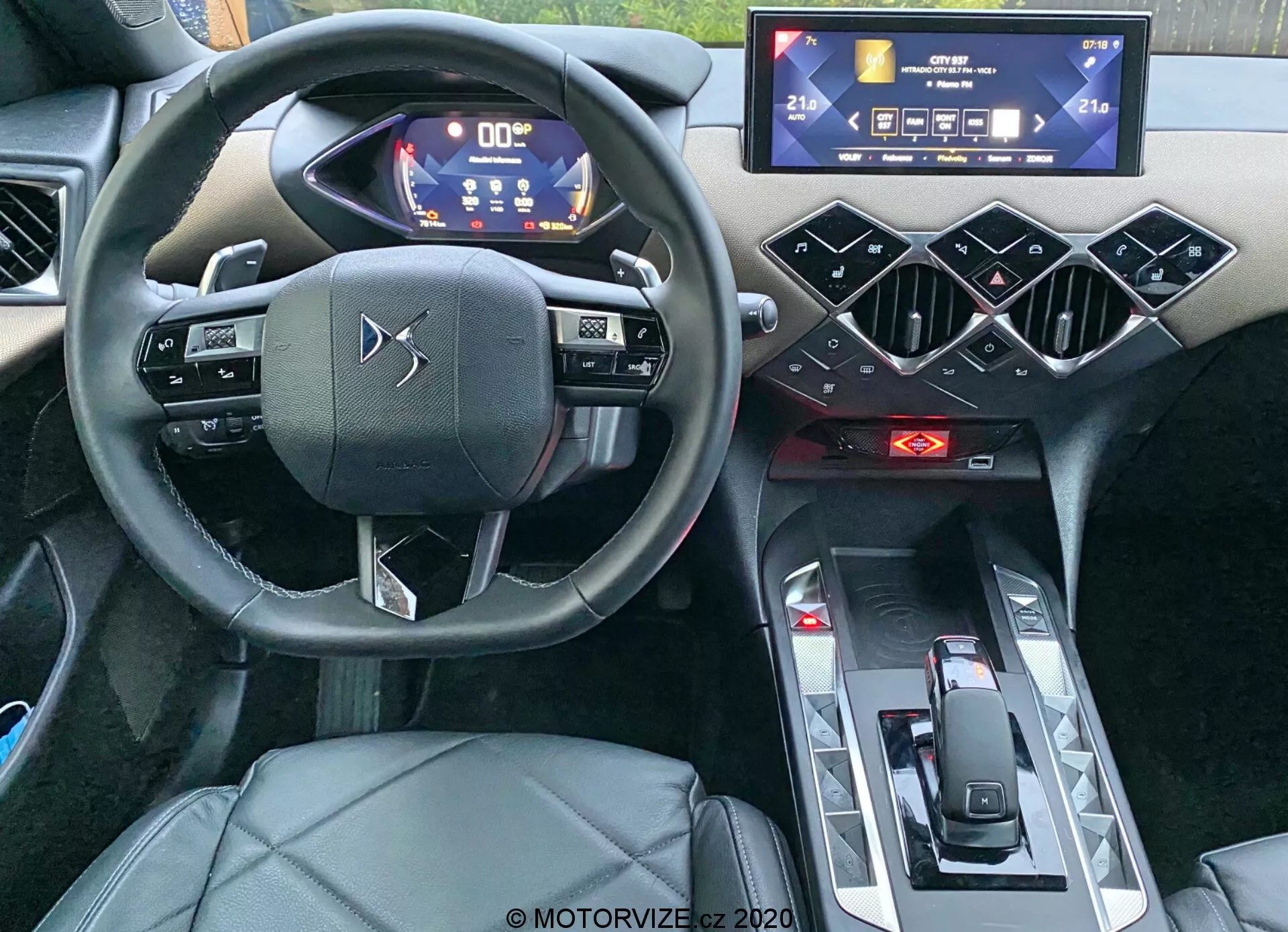 Notranji pogled DS Automobiles DS-3 Crossback 2019, osredotočen na voznikov kokpit iz perspektive višine oči. Posnetek, narejen iz območja zadnjega potnika, poudarja trikraki volan z zmogljivimi dotikalnimi kontrolami, digitalno instrumentno ploščo z modro in belo osvetljenimi merilniki, horizontalno usmerjen zaslon na dotik infotainment sistema, geometrijske šesterokotne oblikovne elemente na sredinski konzoli, rdeče indikacije kontrole klime, minimalističen izbirnik prestav in voznikov sedež oblečen v usnje ali usnju podobno tkanino. Fotografija poudarja sodobne visokotehnološke funkcije in luksuzno oblikovanje avtomobila.