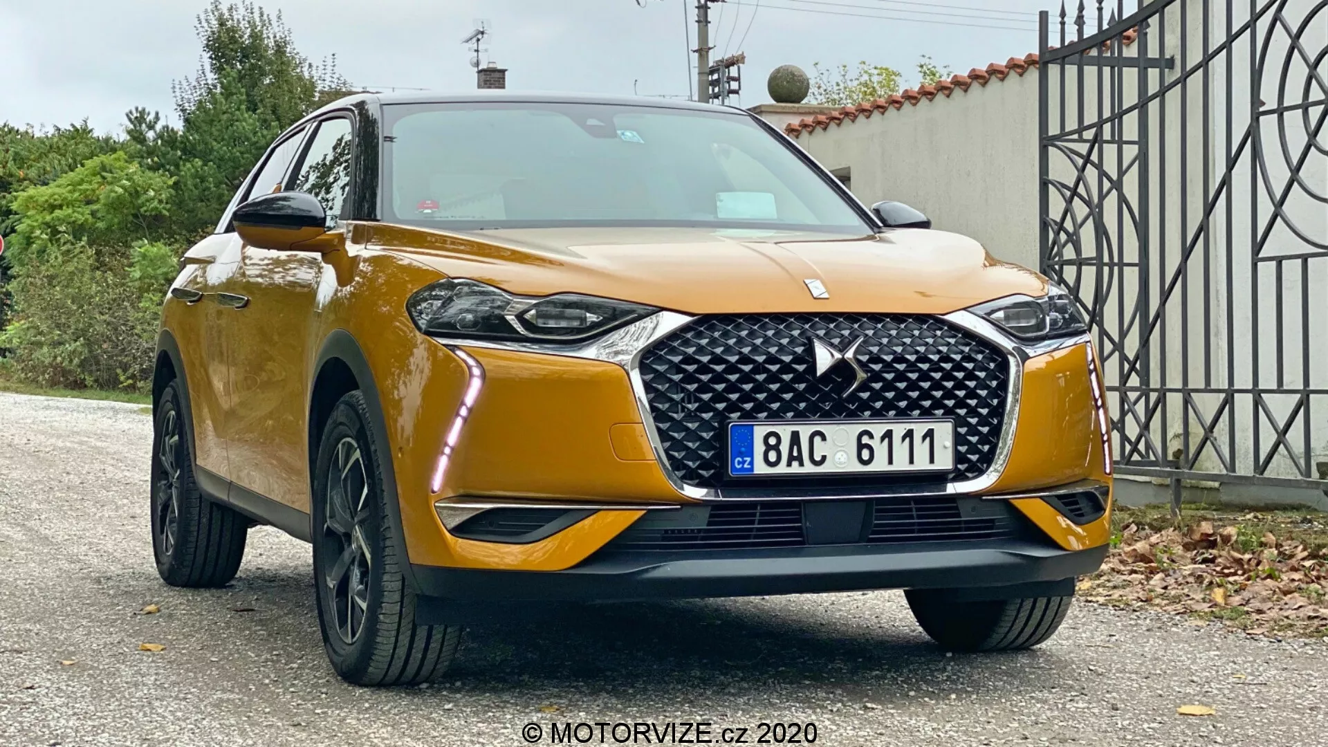 Spredaj trikraten pogled na SUV DS Automobiles DS 3 Crossback 2019, ki prikazuje svojo višino in robustno držo v primestnem okolju. Vidna so kovinsko zlata barva, izrazita mreža hladilnika z diamantnim vzorcem in emblemom, elegantni LED žarometi, meglenke, masiven sprednji odbijač s širokim dovodom zraka, črna večkraka lita platišča in stranska ogledala z vgrajenimi smerniki.
