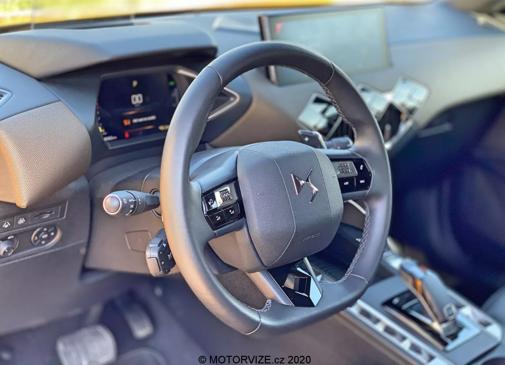 Od blizu notranji pogled DS Automobiles DS-3 2019 (DS-3 Crossback) iz voznikove perspektive, osredotočen na sodoben usnjen volan, ki je nameščen v središču okvirja z večfunkcijskimi kontrolami. Za volanom je digitalna instrumentna plošča, ki je delno ovirana in nakazuje napredno tehnologijo vozila. Spodnji del nakazuje na sredinsko konzolo s prestavno ročico in morebitnimi izbirniki voznih načinov, kar odraža prefinjene notranje in udobne funkcije.