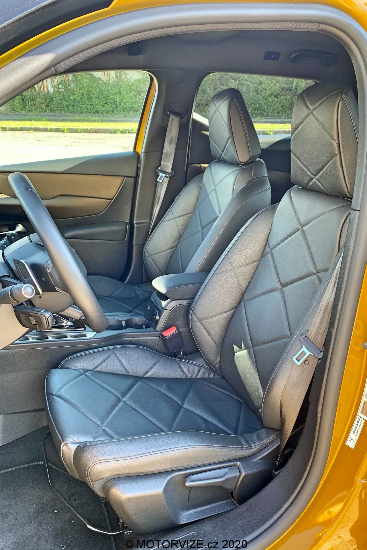 Pogled v notranjost DS Automobiles DS-3 Crossback 2019, osredotočen na sprednje sedeže z vidika sopotnikovih vrat. Slika poudarja črne usnjene prešite sedeže z vgrajenimi vzglavniki in stranskimi oporami, kar nakazuje luksuzen ali športen dizajn. Vidni so del sredinske konzole z prestavno ročico, ki nakazuje samodejni menjalnik, ter spodnji del armaturne plošče. Notranja plošča vrat ustreza usnju sedežev, s kovinskim ročajem vrat in kontrolnimi gumbi. Vidni so tudi črni varnostni pasovi z mehanizmom za nastavitev višine, preproga na tleh in kontrastna zunanja barvna shema, verjetno rumena.