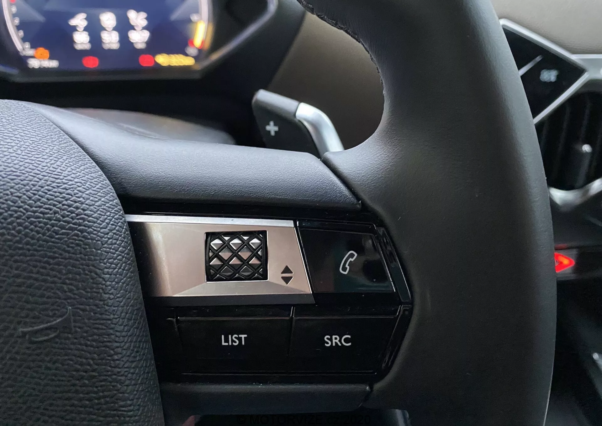 Od blizu iz voznikove perspektive na levi strani volana v DS Automobiles DS 3 Crossback 2019. Sliko se osredotoča na kontrolne gumbe, vključno z gumbom s telefonsko ikono, gumbom za 'SEZNAM' in gumbom za 'SRC', poudarjenimi na zamegljenem ozadju armaturne plošče. Visoko kontrastna zasnova združuje črne in kovinske obloge z diamantnim vzorcem na kromu, kar poudarja njegov vrhunski videz.
