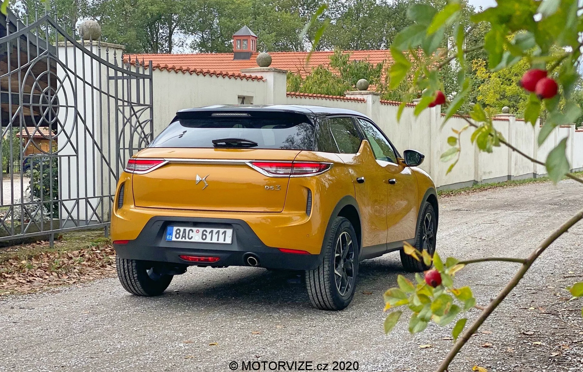 Pogled od zadaj pod kotom na DS Automobiles DS 3 Crossback 2019, zajet iz nizke perspektive na stanovanjskem dovozu. Avto ima izrazite horizontalno stilizirane zadnje luči, centralno nameščen logotip proizvajalca na skulpturiranih prtljažnih vratih z integriranim spojlerjem in tretjo zavorno lučjo, centralno nameščeno registrsko tablico in sijoč kovinski oranžni ali zlat odtenek. Robustni zadnji odbijač ima vgrajene senzorje ali odsevnike, z vidnim zadnjim brisalcem, anteno v obliki plavuti morskega psa in večkrakimi litimi kolesi. Malce zamegljena vegetacija v ospredju kontrastira z ostrejšo osredotočenostjo na vozilo in vhodnimi vrati v ozadju, kar nakazuje mehko razpršeno dnevno svetlobo.