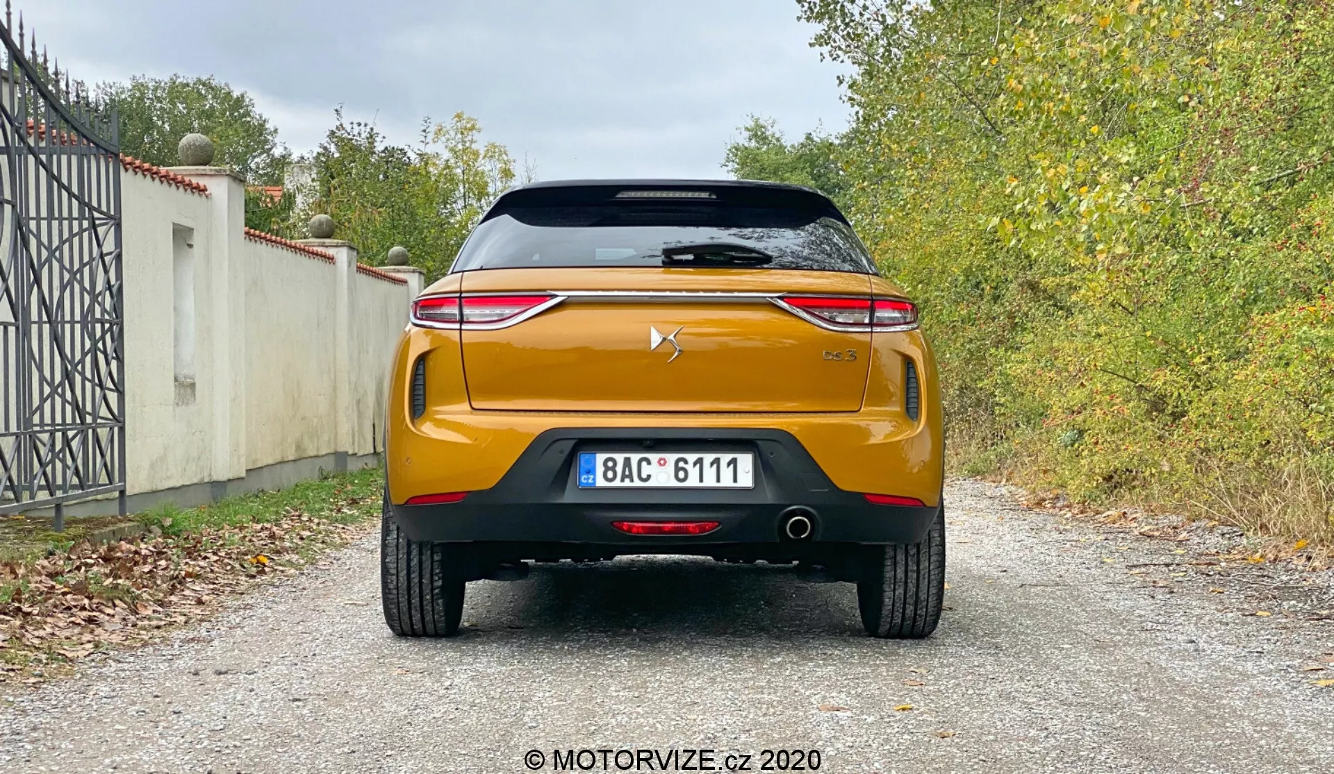 Pogled zadaj na moderni avto DS-Automobiles DS-3 Crossback 2019, ki prikazuje njegovo obliko in tehnološke lastnosti, vključno s strešnim spojlerjem, zadnjim vetrobranskim steklom z grelnim elementom, visoko nameščeno zavorno lučjo, pokrovom prtljažnika z logotipom proizvajalca, izstopajočim kromiranim trakom, elegantnimi LED zadnjimi lučmi, robustnim zadnjim odbijačem s parkirnimi senzorji, dvojnim izpušnim sistemom, črnim difuzorskim elementom in presenetljivo oranžno ali bakreno barvo na gramozni površini z vegetacijo v ozadju.