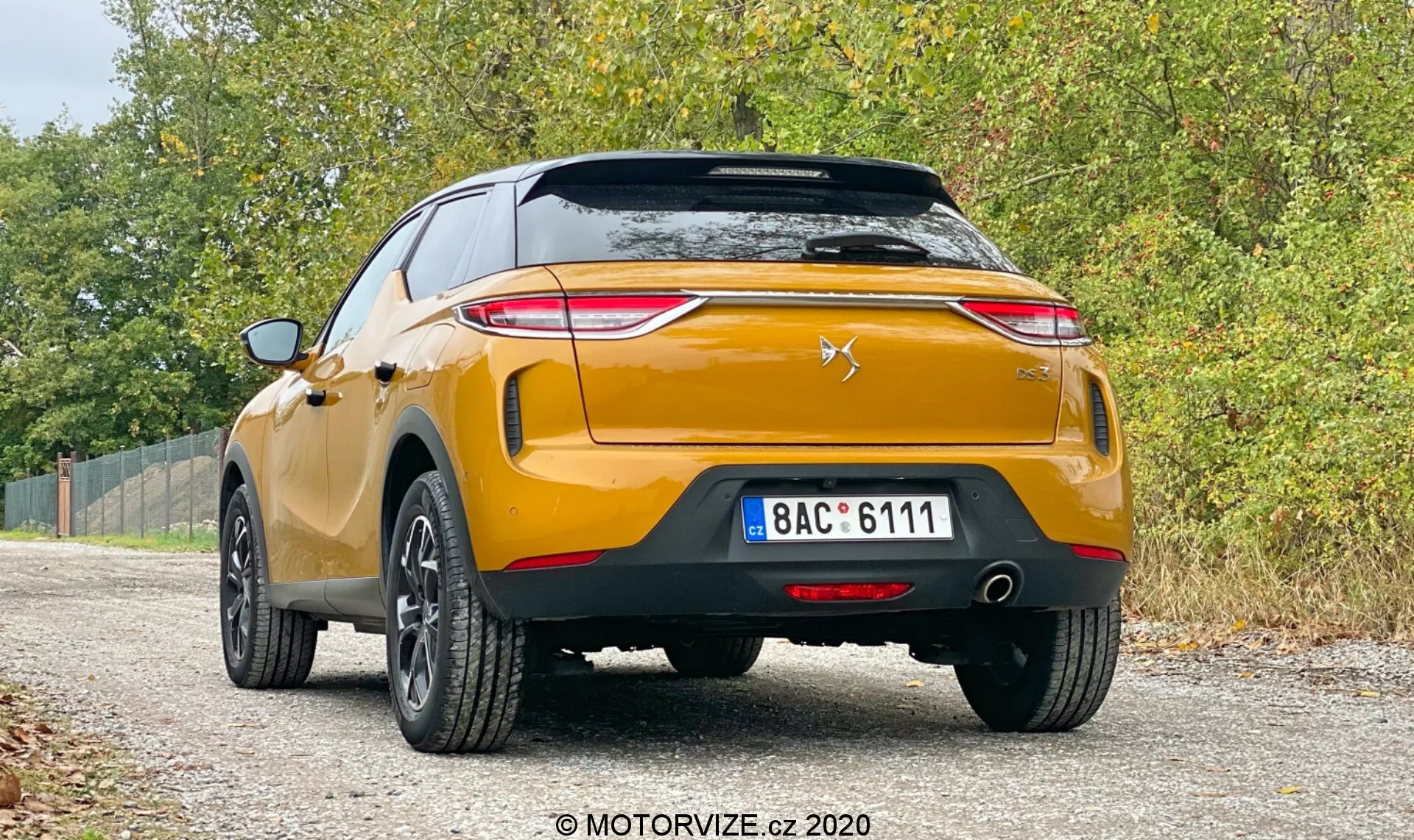 Slika prikazuje zadnji pogled na moderen, zlat DS-3 Crossback 2019, parkiran na neasfaltirani cesti z vegetacijo na obeh straneh. Avto, posnet z nizkega kota, prikazuje ostre zadnje luči, usmerjene proti sredini prtljažnika, stiliziran emblem v obliki chevrona z levjim silhuetom na prtljažniku, robusten zadnji odbijač s parkirnimi senzorji, dvojne izpušne cevi, črno plastično obrobo in pet-kraka platišča iz lahke zlitine. Registrska tablica je evropskega sloga, s standardnimi zadnjimi odbojniki in zadnjo meglenko. Mehka, razpršena osvetlitev nakazuje oblačen dan, kar prispeva k priložnostnemu, vsestranskemu ozračju, medtem ko vodni žig ‘MOTORVIZE.cz 2020’ kaže na vir slike.