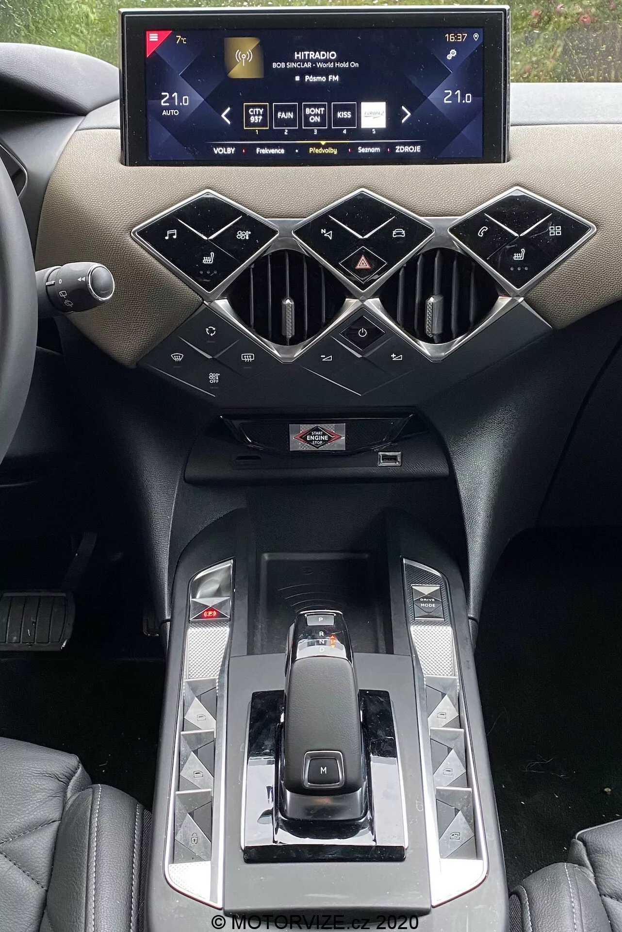 Notranjost DS Automobiles DS 3 2019 s poudarkom na sredinski konzoli z infotainment sistemom. Širok, horizontalno usmerjen zaslon na dotik prikazuje nastavitve vozila, vključno z nastavitvami temperature klime, nastavljeno na 21,0 °C. Spodaj so geometrično oblikovane prezračevalne odprtine in fizični gumbi v obliki diamanta za upravljanje sistemov avtomobila. Moderen prestavni ročaj, sijoče plošče in namigovanje na držala za pijačo so poudarjeni z luksuznimi materiali in premium oblikovanjem.