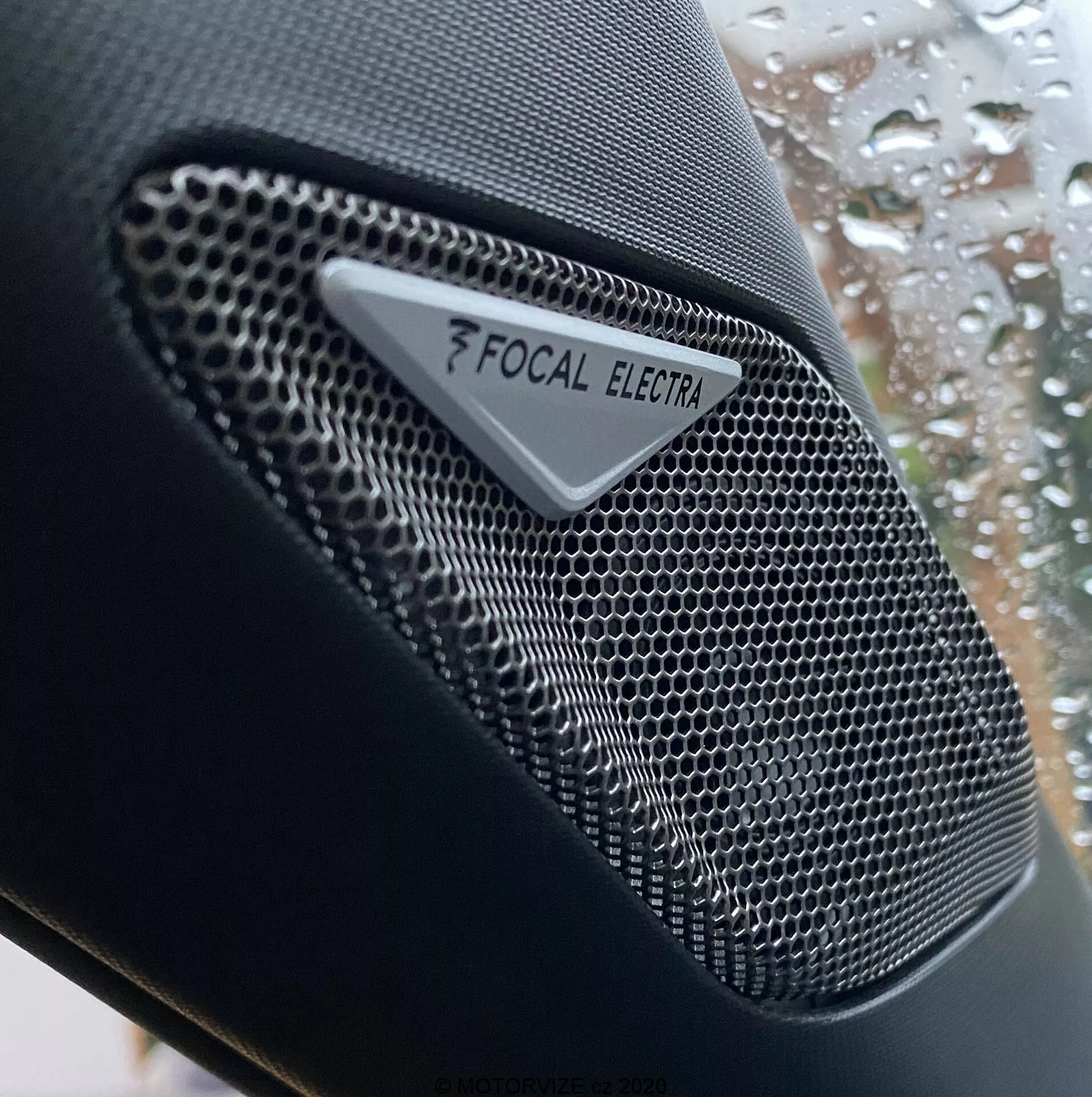 Pogled od blizu na zvočnik s Focal Electra blagovno znamko v DS Automobiles DS-3 Crossback 2019, ki prikazuje visokofrekvenčno avdio komponento s kovinsko mrežo v obliki satovnice in jasnim logotipom Focal ter blagovno znamko Electra. Posnetek se osredotoča na zapleteno zasnovo zvočnika, s plitko globinsko ostrino, ki poudarja teksturo zvočnika v primerjavi z zamegljenim ozadjem avtomobilskega okna z dežnimi kapljicami.