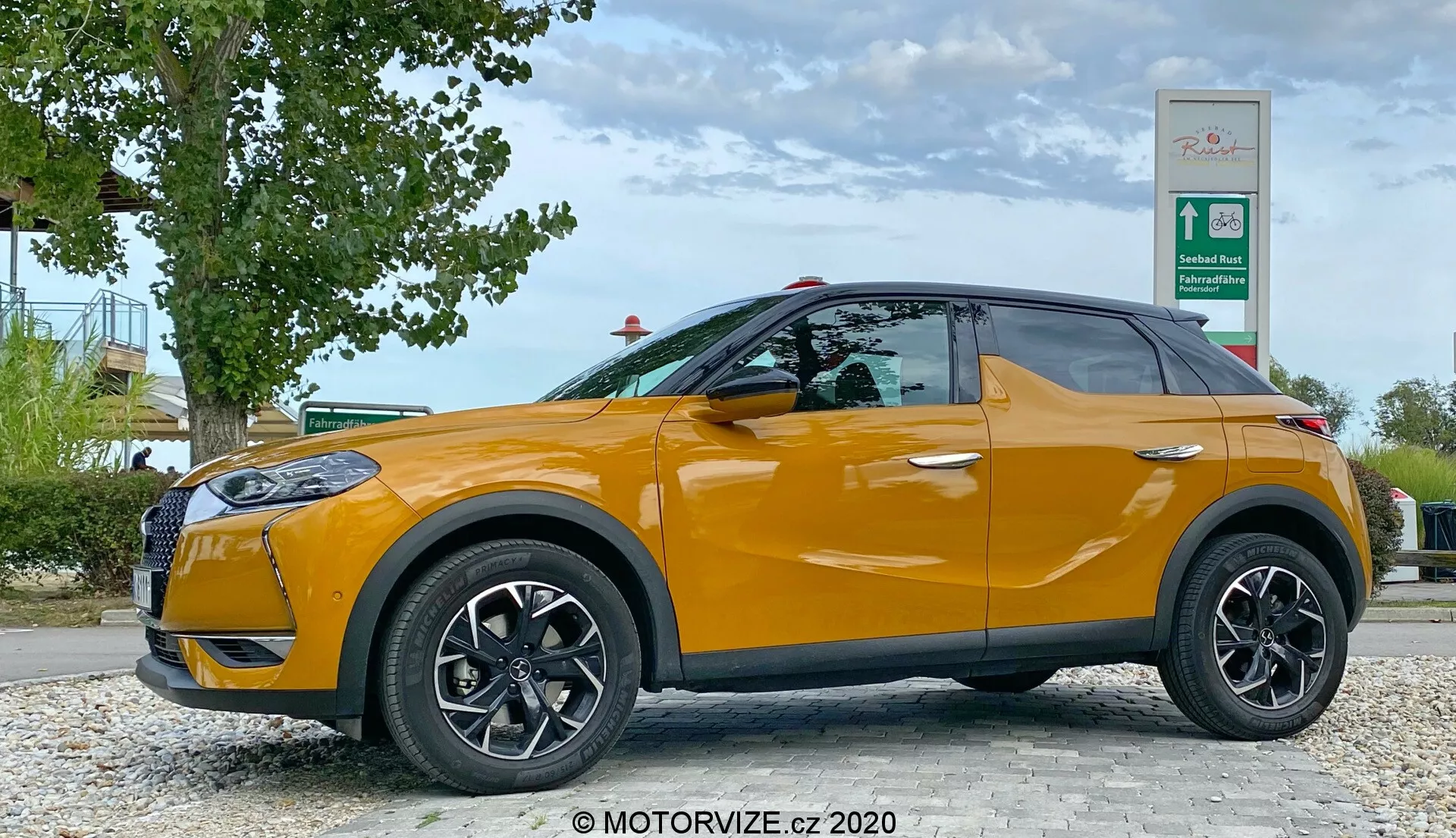 Stranski profil DS Automobiles DS 3 Crossback 2019 v svetlo oranžni barvi, parkiran ob cesti z zelenjem in znakom za bencinsko črpalko v ozadju. Voznikova stran z črnimi večkrakimi kolesi in stranskimi ogledali v barvi karoserije je vidna iz nizkega kota na višini koles avtomobila, prikazuje elegantno obliko kompaktnega SUV-ja s dvigajočo se pasno linijo in zatemnjenimi okni.