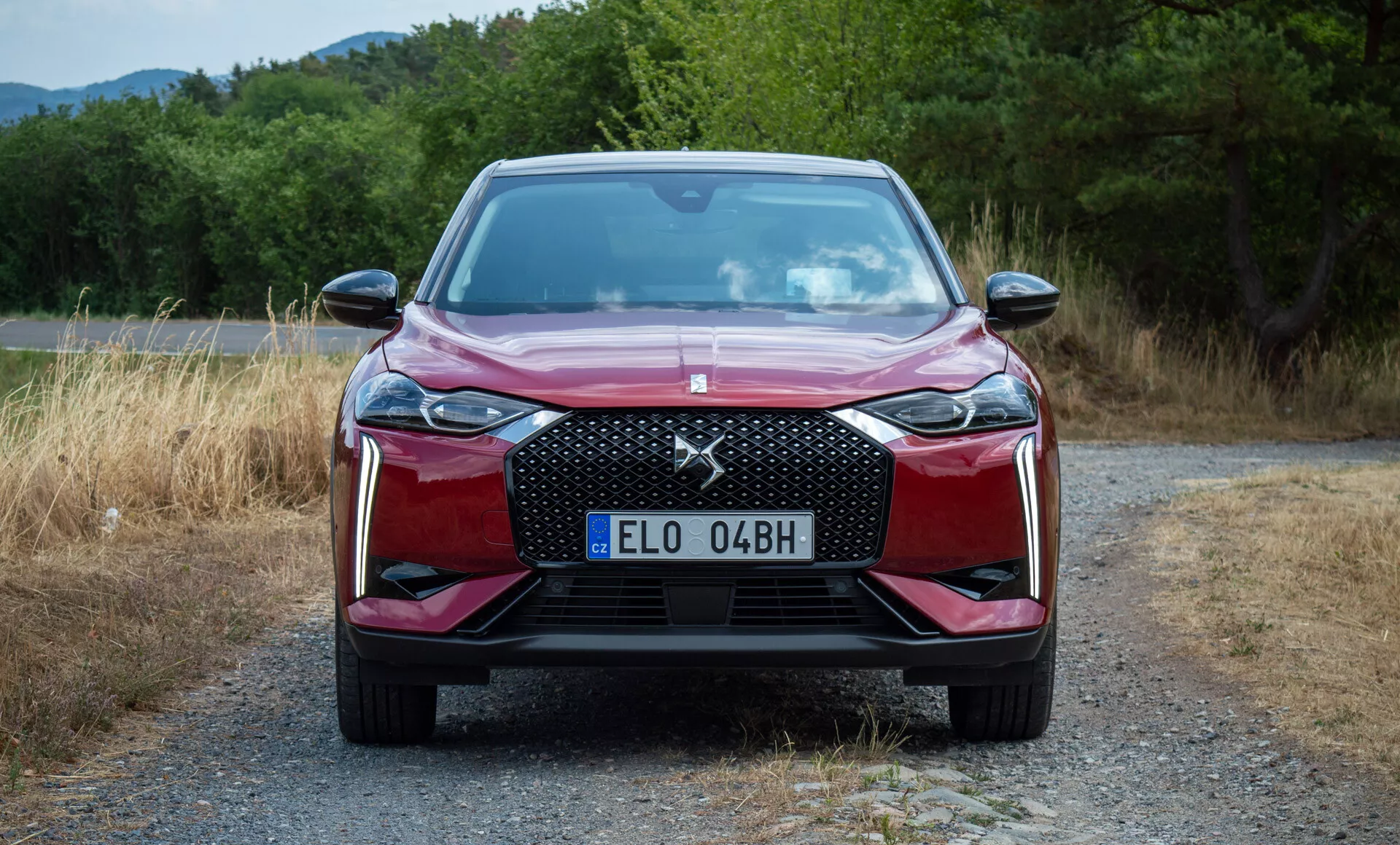 Sprednji pogled na DS Automobiles DS 3 E-TENSE 2023, parkiran na gramozni poti z zelenjem v ozadju, predstavlja sprednjo rešetko, LED žaromete, linije pokrova in zasnovo odbijača