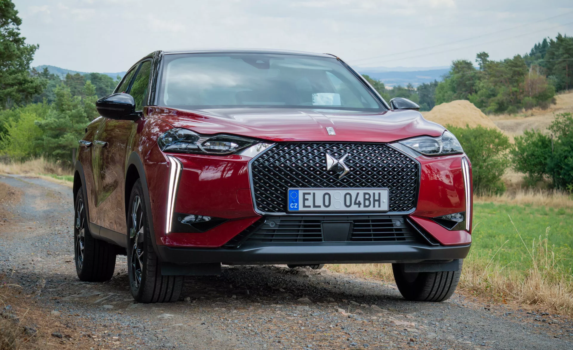 Moderen rdeč avtomobil DS Automobiles DS 3 E-TENSE 2023, zajet z nizkega sprednjega kota, ki poudarja sprednjo rešetko, emblem in delno bočni profil na makadamski cesti z naravnim ozadjem