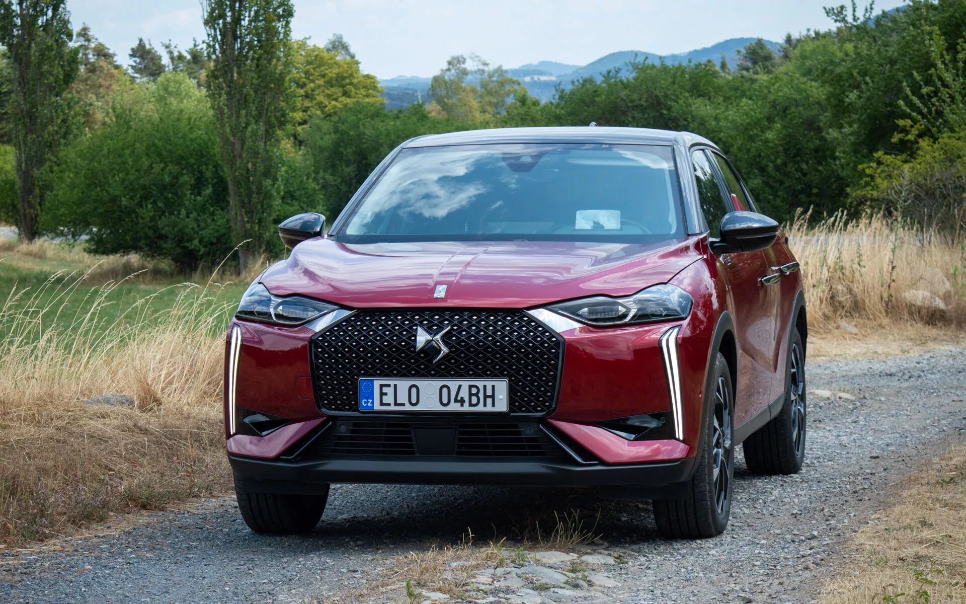DS Automobiles DS 3 E-TENSE leta 2023 prikazan na gramozni poti, ki prikazuje svojo šesterokotno rešetko, LED žaromete, sofisticiran sprednji odbijač in metalno rdečo barvo pred naravnim ozadjem z drevesi in travo