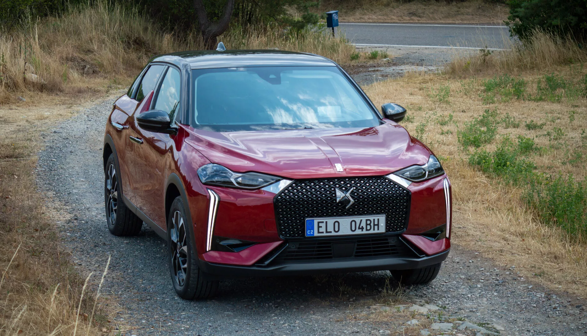 DS Automobiles DS-3 E-TENSE 2023 v metalik rdeči, prikazuje svoj sodoben pogled spredaj tričetrt, vključno z detajlno rešetko, LED žarometi in aerodinamično obliko karoserije, parkiran na podeželski cesti, kar nakazuje vsestranskost