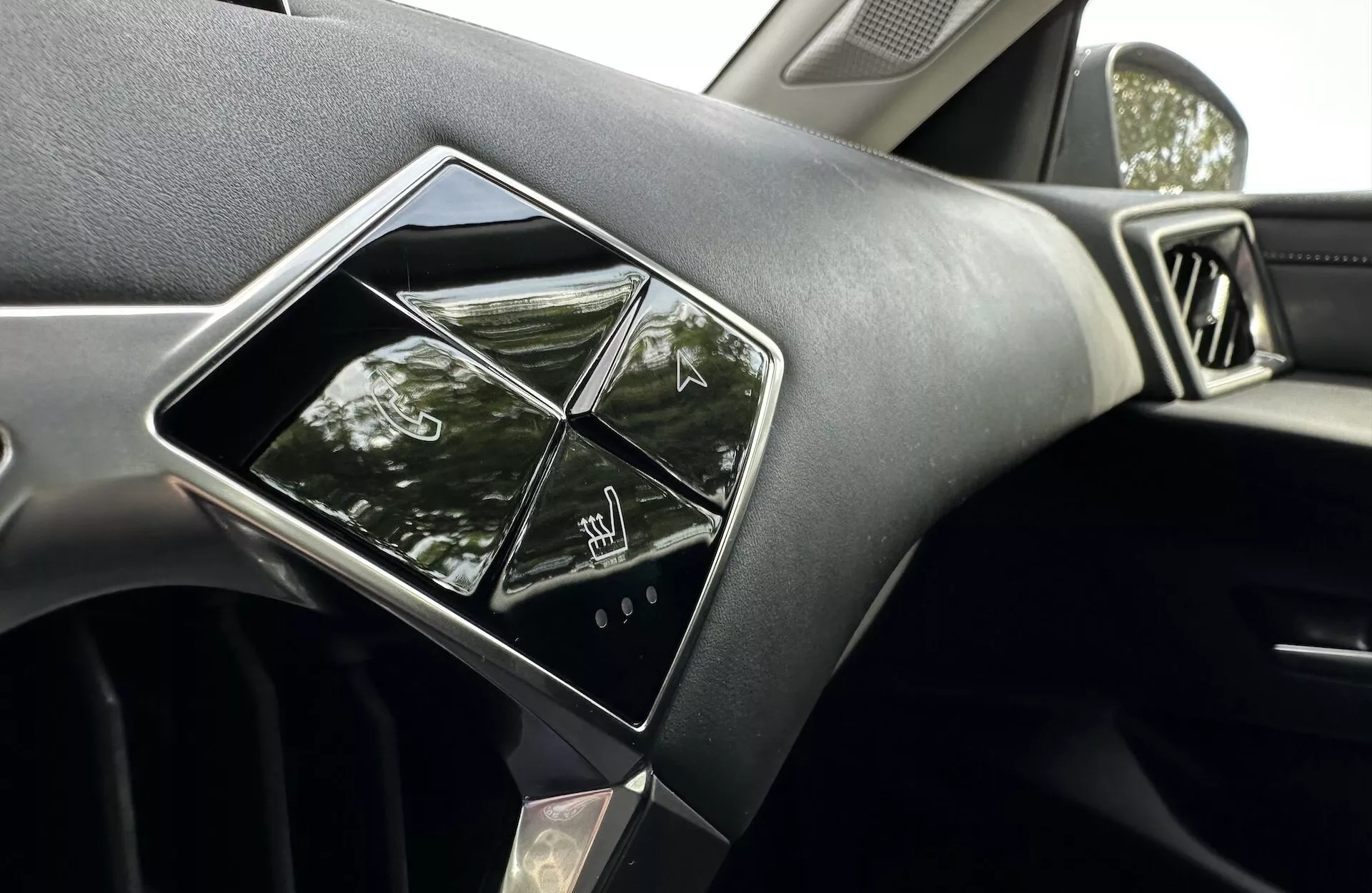 Pogled na notranjost DS 3 E-TENSE 2023, osredotočen na armaturno ploščo in sistem za infotainment, z značilnimi občutljivimi kontrolniki na dotik, premium dizajnom armaturne plošče z materialom, ki je mehek na dotik ali podoben usnju, delom zračnika in odsevi, ki kažejo na naravno dnevno svetlobo in zunanje parkirno okolje.