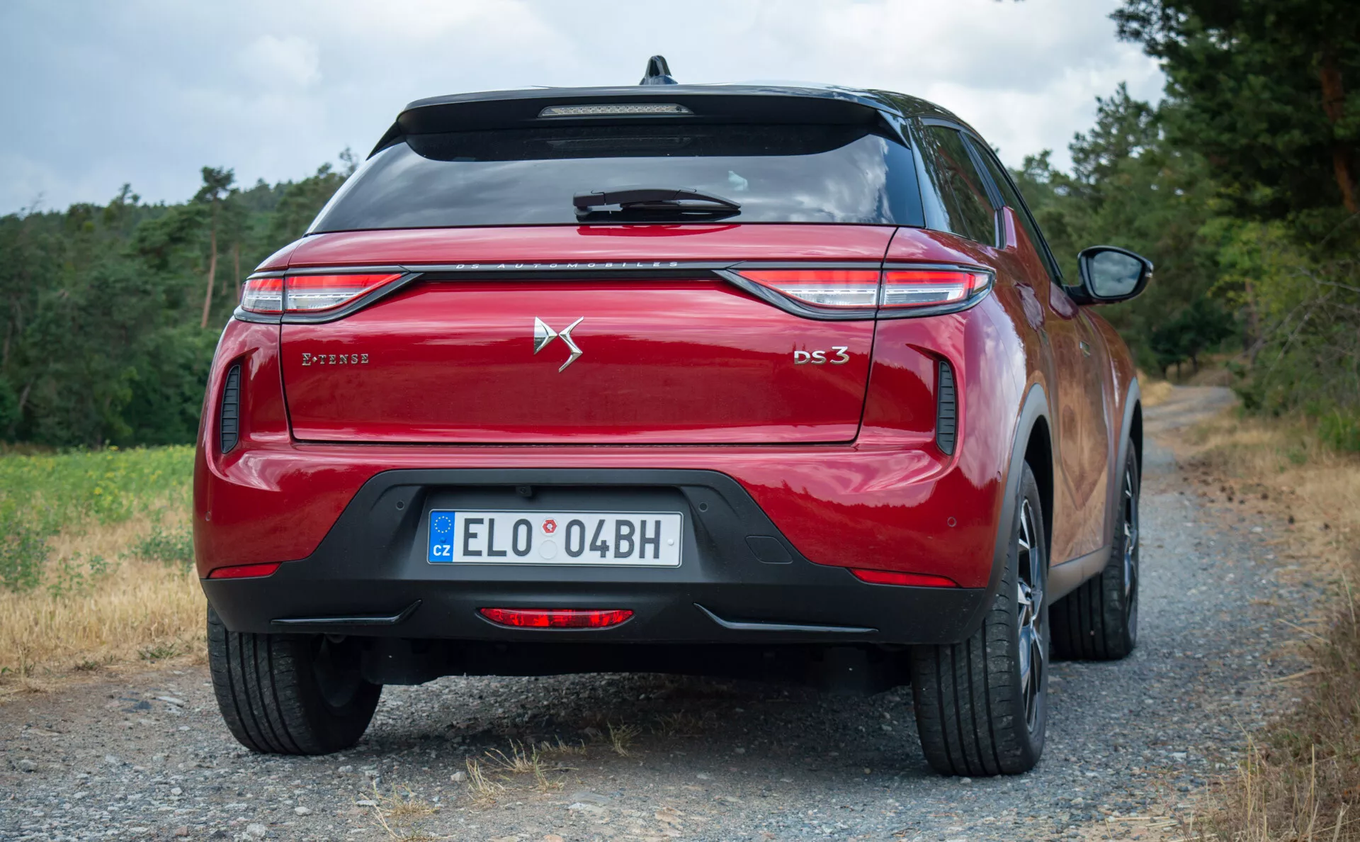 Zadnji pogled na rdeč model DS 3 E-TENSE 2023, ki prikazuje njegove moderne zadnje luči, dvojne izpušne cevi in značko, postavljeno na prostem na gramozni cesti z vegetacijo.