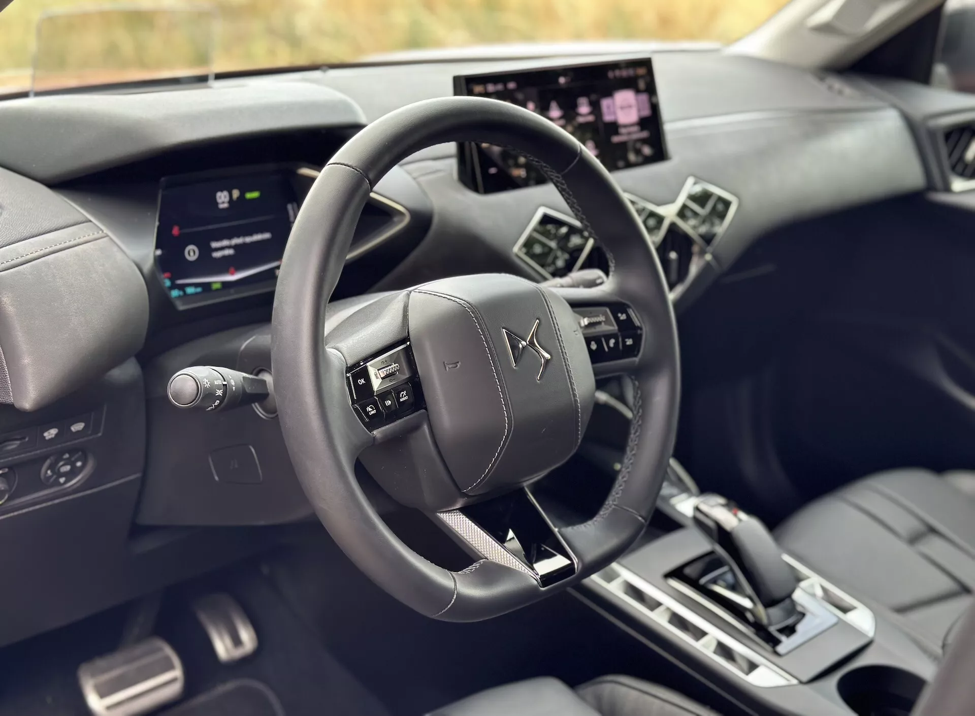 Pogled na notranjost DS 3 E-TENSE 2023 iz perspektive voznika - osrednji volan oblečen v usnje z emblemom DS Automobiles, digitalni instrumentni sklop in velik zaslon na dotik infotainment sistema med razkošnimi temnimi toni z angularnimi oblikovnimi linijami.