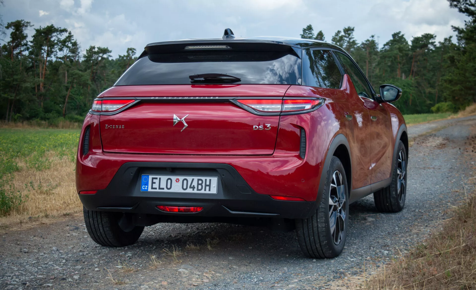 Hatchback DS Automobiles DS 3 E-Tense 2023 v kovinski rdeči barvi, pogled od zadaj zgoraj, ki poudarja zadnje luči, emblem in oznako modela, postavljen na makadamski cesti z naravnim ozadjem.