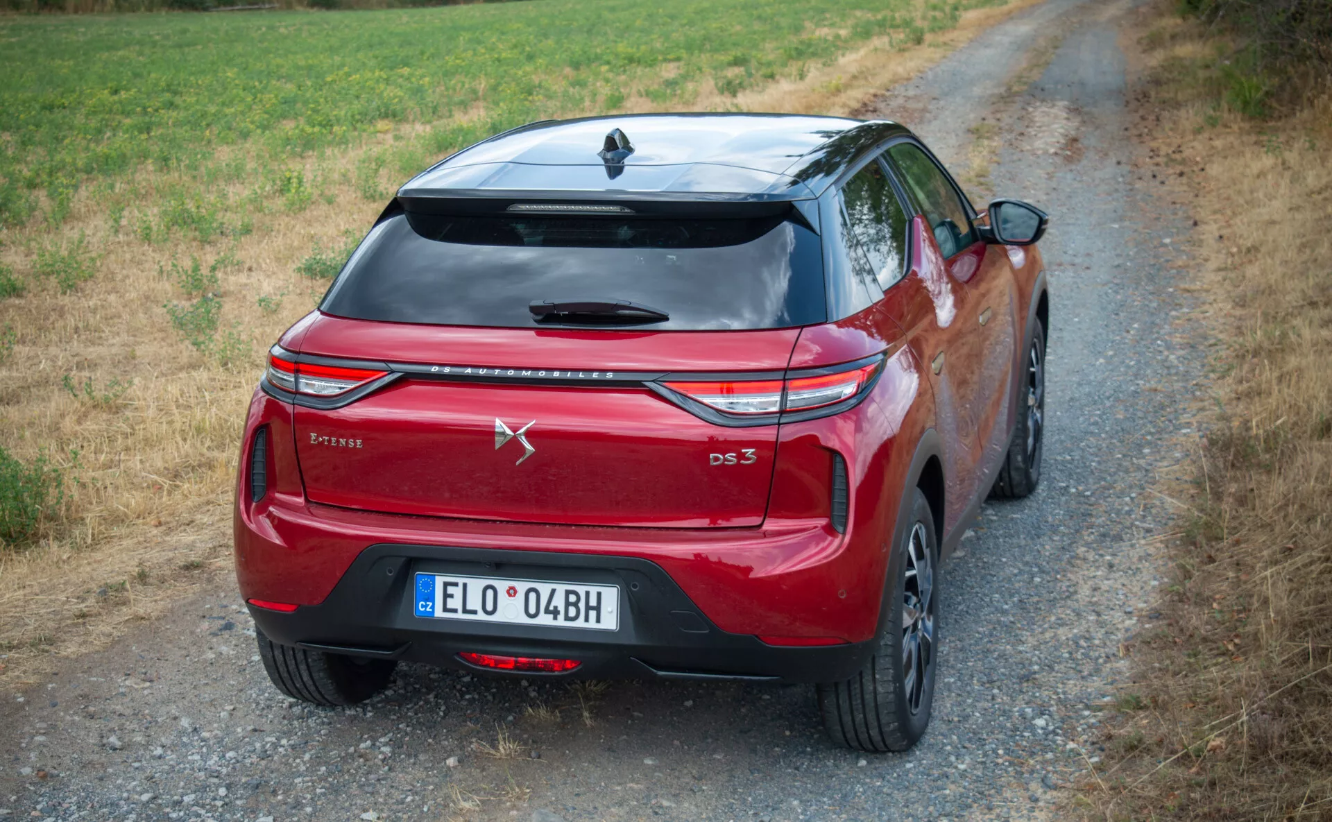 Sodoben kompakten SUV DS Automobiles DS-3 E-TENSE 2023 parkiran na makadamski cesti obdan z naravo, prikazuje LED zadnje luči, oznako CROSSBACK, stilsko oblikovana vrata prtljažnika s spojlerjem, športna lita platišča in anteno v obliki morskega psa v ruralnem okolju