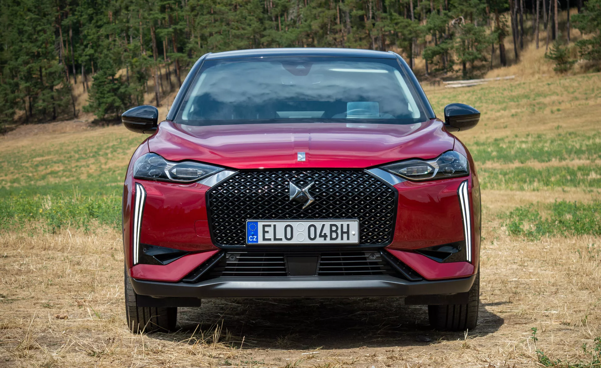 DS Automobiles DS 3 E-TENSE 2023 v rdeči barvi, ki prikazuje značilne lastnosti iz sprednjega kota na neurejeni površini z gozdnim ozadjem, osvetlitev sredi dne poudarja njegovo teksturirano rešetko, LED žaromete, aerodinamični odbijač z zračnimi vstopi, evropsko registrsko tablico in sijočo barvo laka.