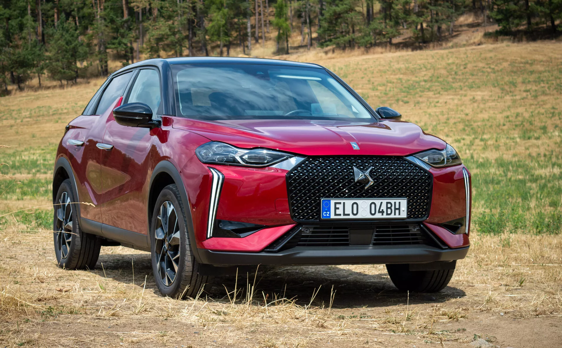Moderen SUV DS Automobiles DS-3 E-TENSE 2023, pogled od spredaj in s strani, ki prikazuje rešetko, pokrov motorja, žaromete in stranski profil na suhi travi z naravnim ozadjem